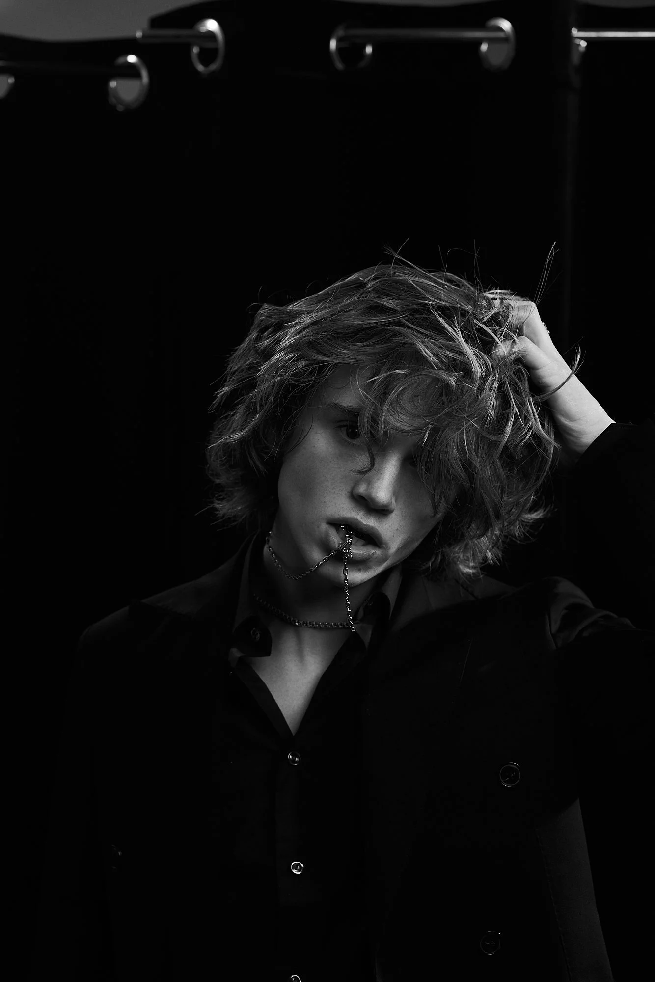 Black and white photo of a young person with tousled hair, wearing a dark jacket and shirt, holding a chain in their mouth, with one hand in their hair, against a dark background.