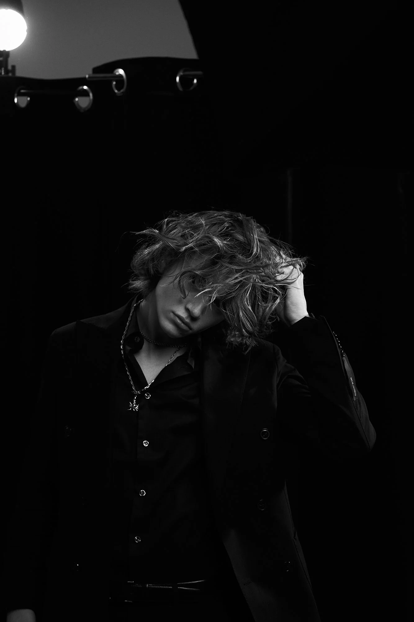 A person with wavy hair, wearing a dark shirt and jacket, poses with one hand in their hair against a dark background, in black and white.