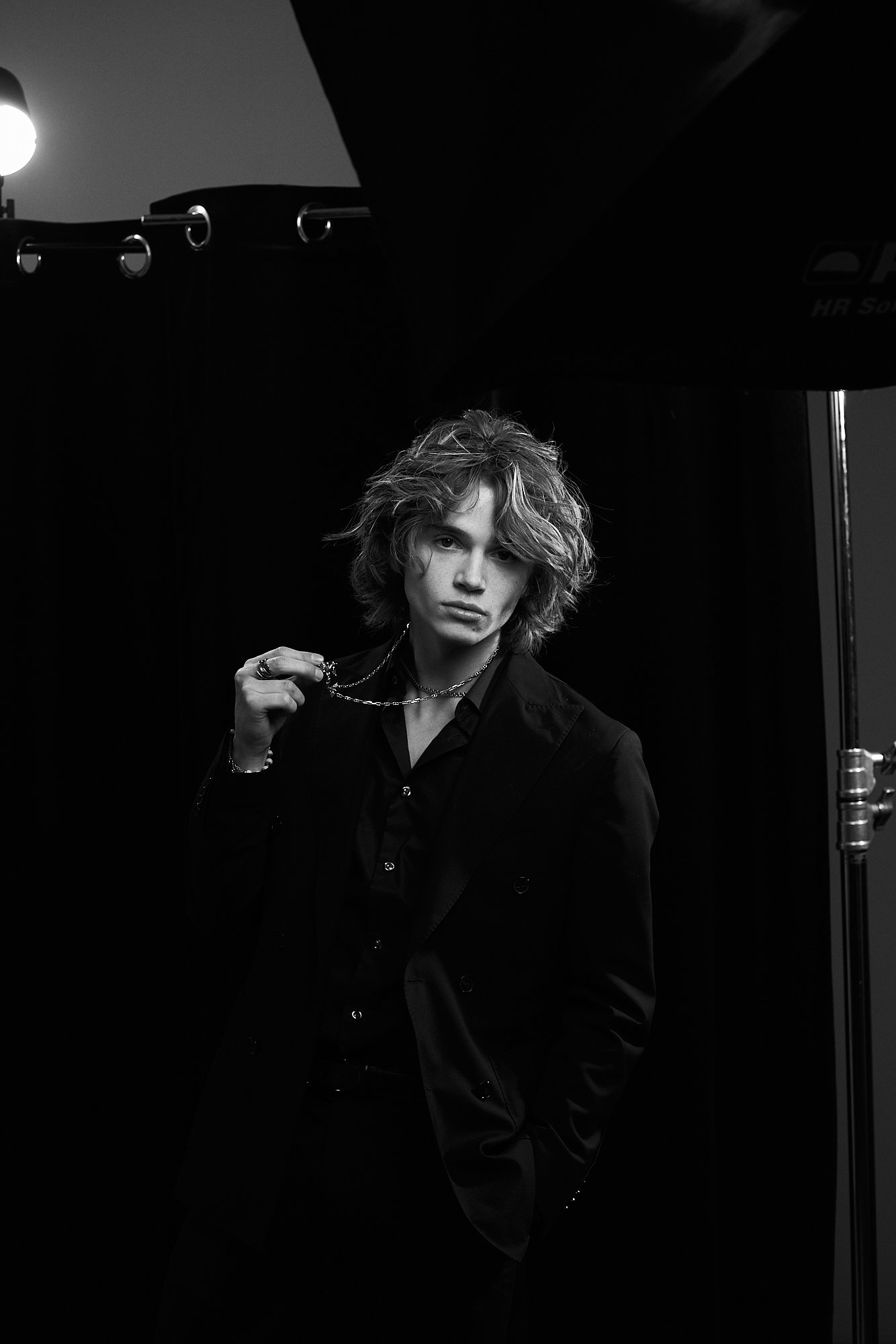 A young man with shoulder-length wavy hair, dressed in a dark suit and shirt, standing in a photography studio with black backdrop and lighting equipment, holding a chain necklace.