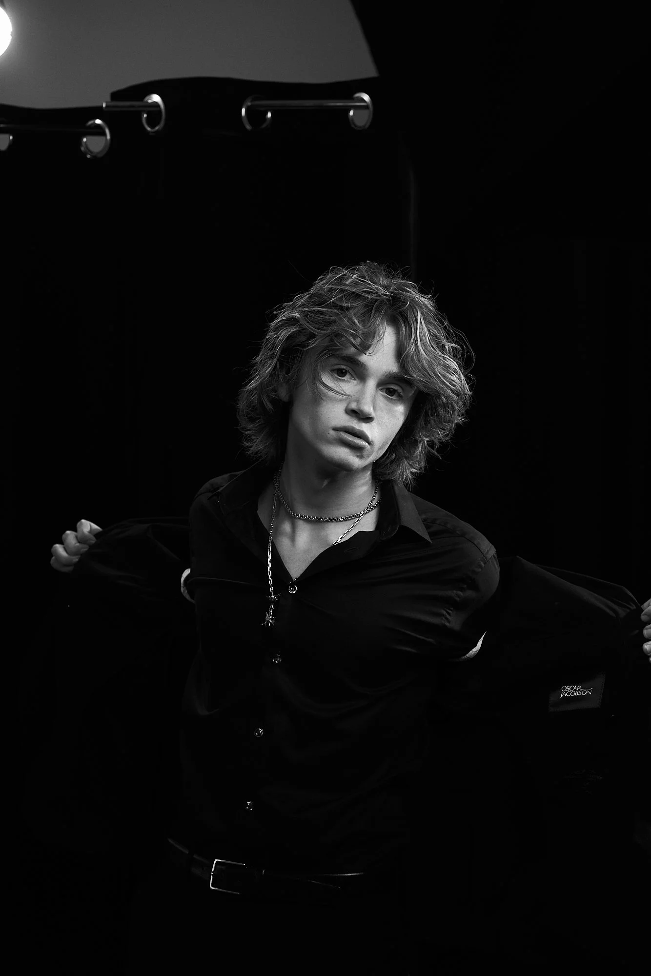 Black and white portrait of a person with wavy hair, wearing a dark shirt, and holding an opened jacket, in front of a dark background.