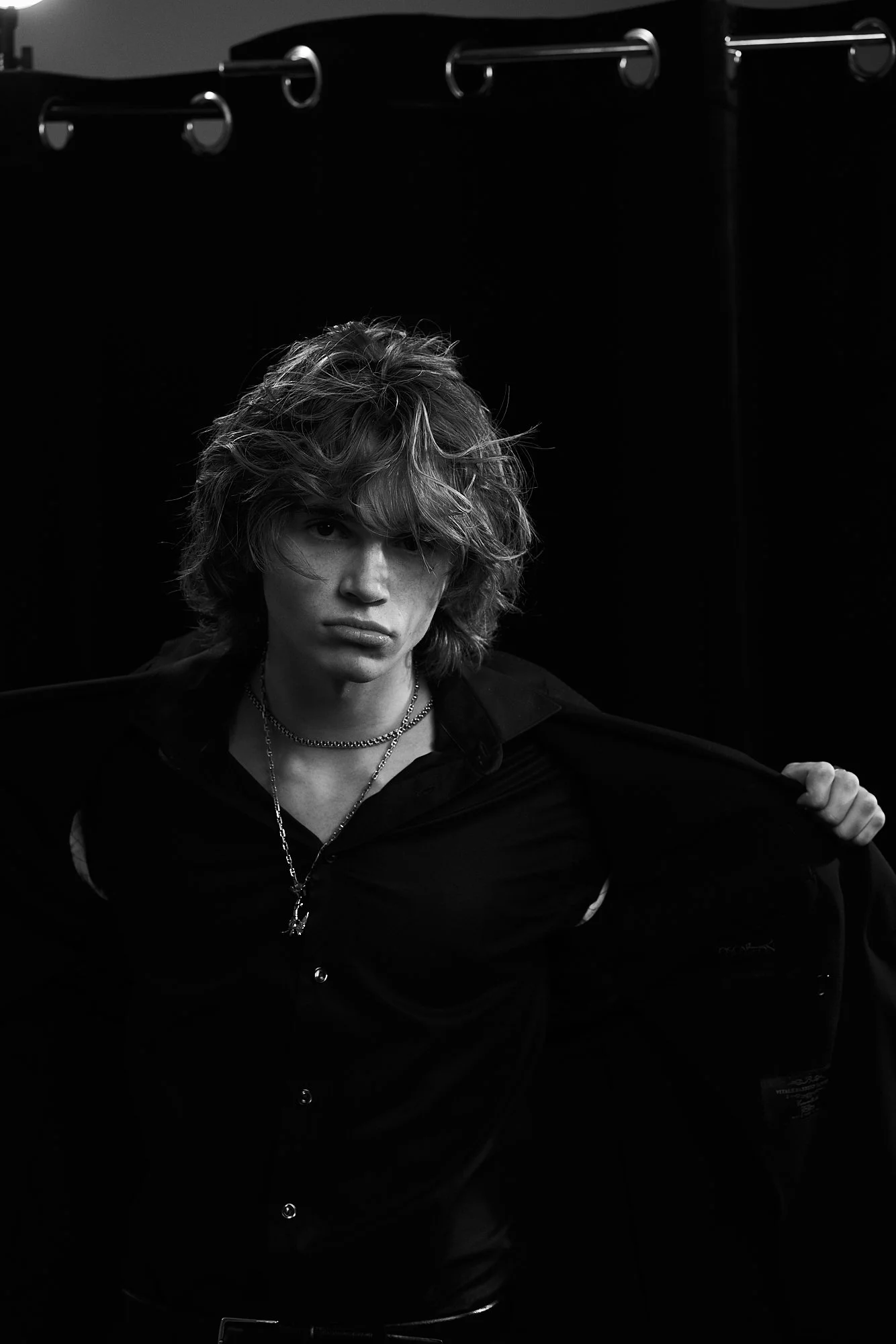 Black and white photo of a young person with wavy, tousled hair, wearing a dark button-up shirt, layered necklaces, and holding an open jacket over their shoulder, set against a dark background.