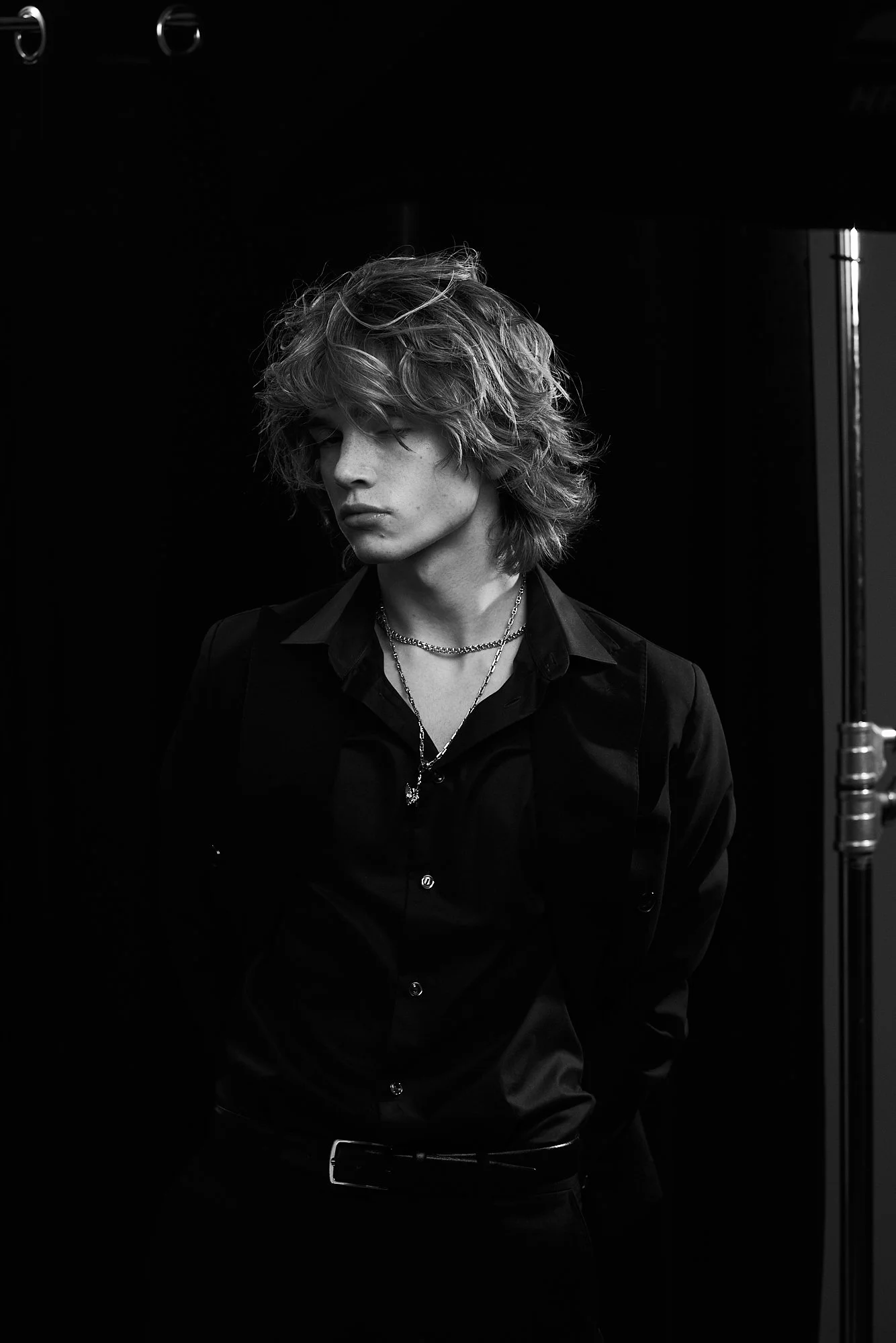 Black and white portrait of a young man with wavy, shoulder-length hair, wearing a dark shirt, layered necklaces, and a belt, against a dark background.