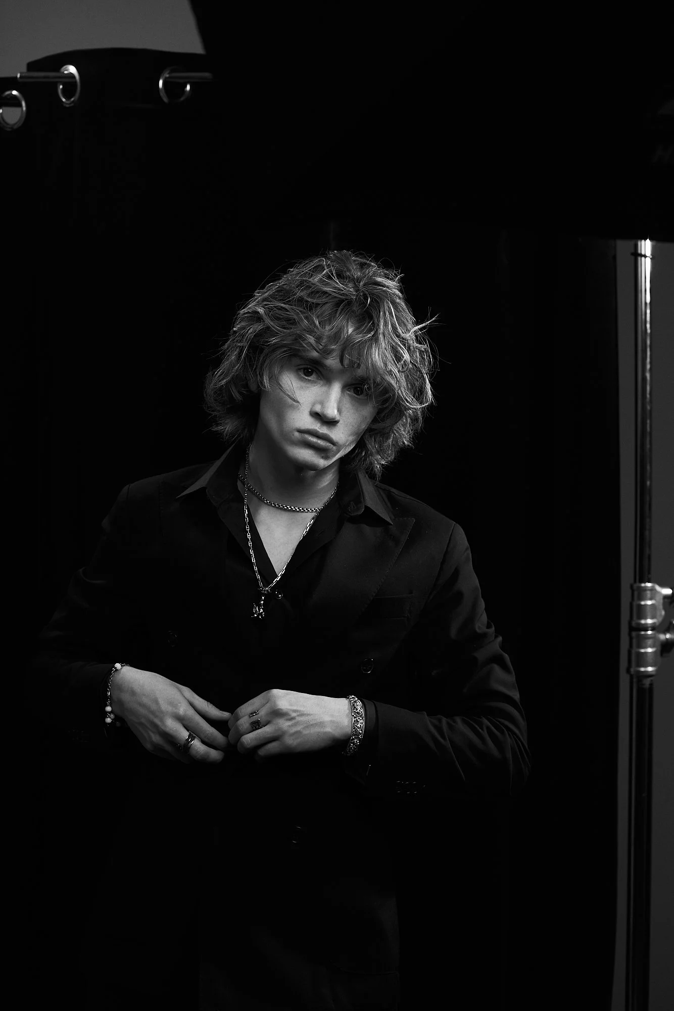 Black and white portrait of a young man with curly hair, wearing a dark suit, layered necklaces, and jewelry, standing against a dark background.