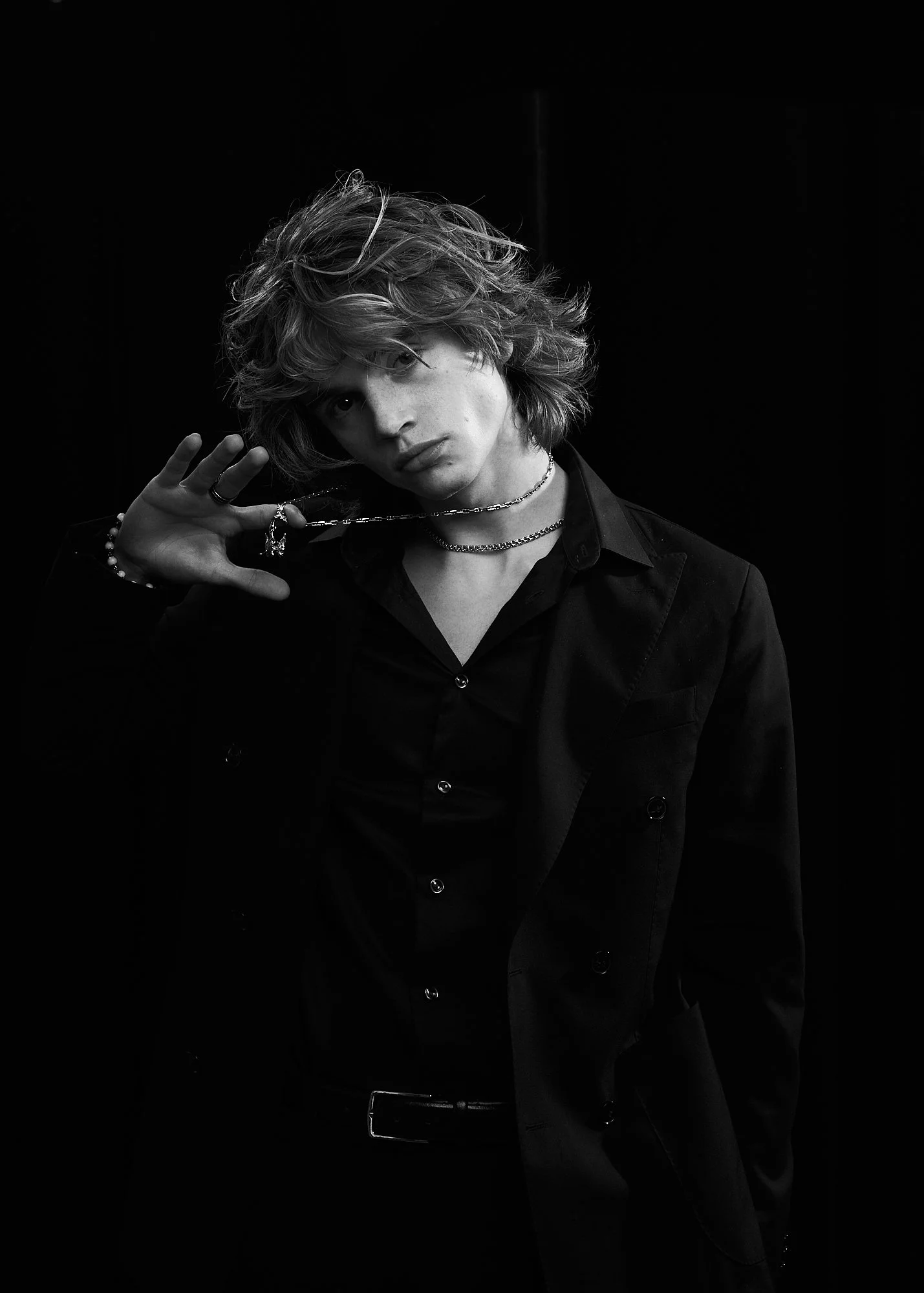 Black and white portrait of a person with wavy hair, wearing a dark suit, jewelry, and posing with one hand near their face against a dark background.