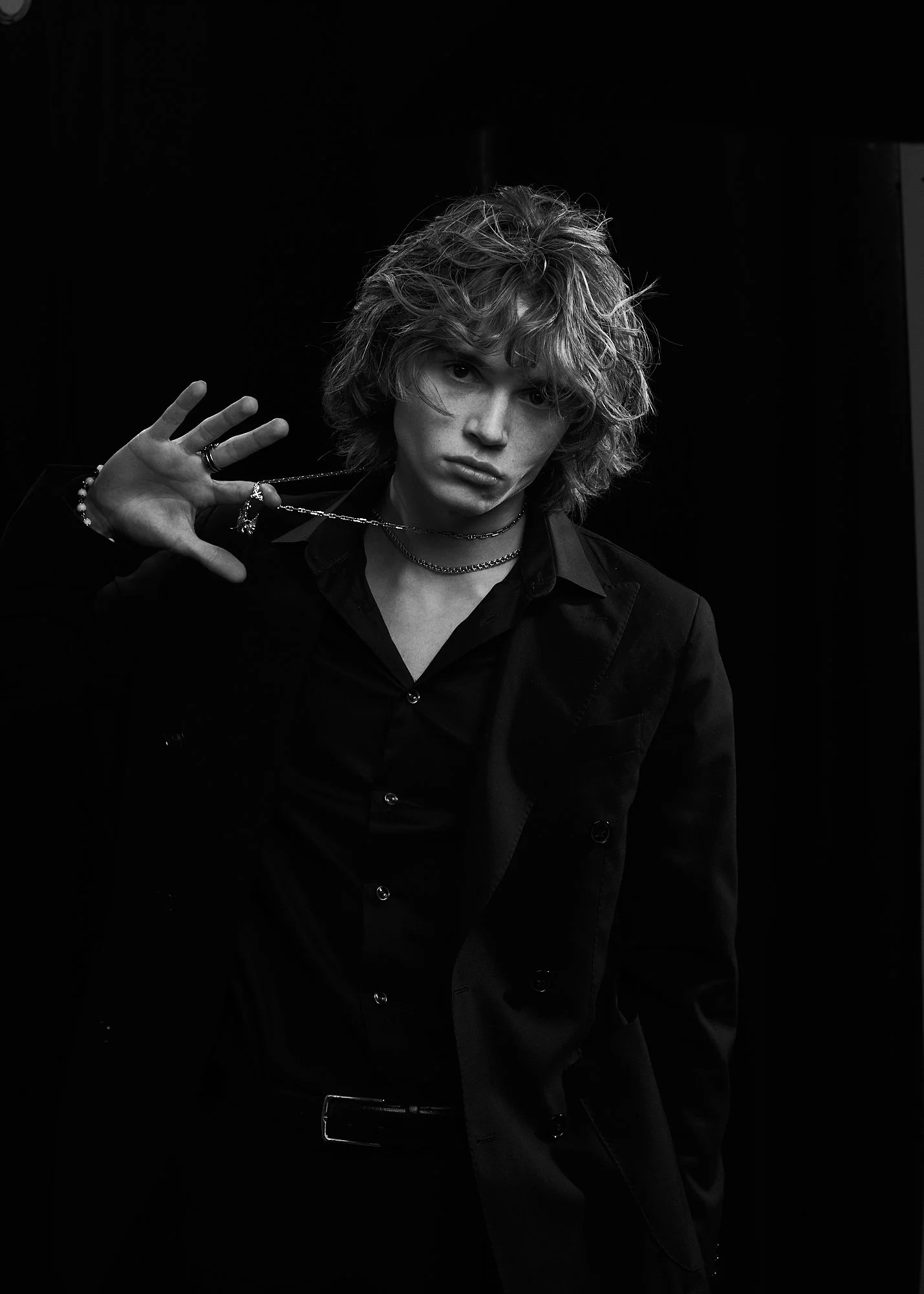 Black-and-white portrait of a young man with curly hair, wearing a dark suit and jewelry, making a gesture with his hand near his face.