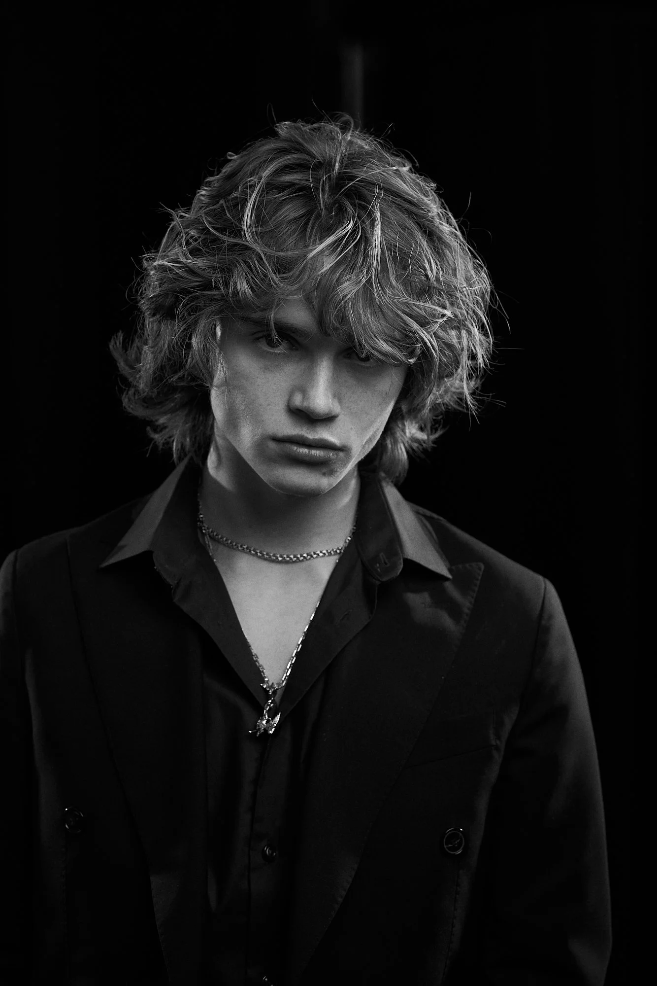Black and white portrait of a young person with wavy, shoulder-length hair, wearing a dark blazer and layered necklaces.