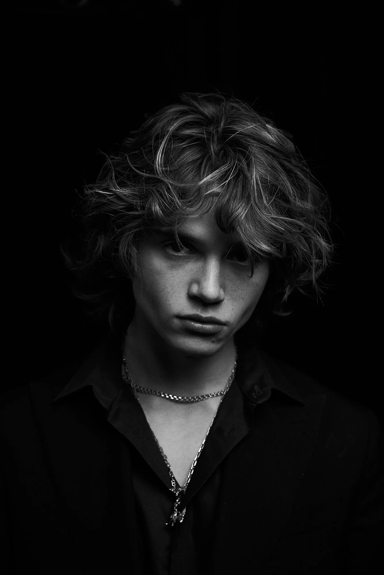 Black and white portrait of a young person with curly, tousled hair, wearing a dark shirt and layered necklaces, against a dark background.