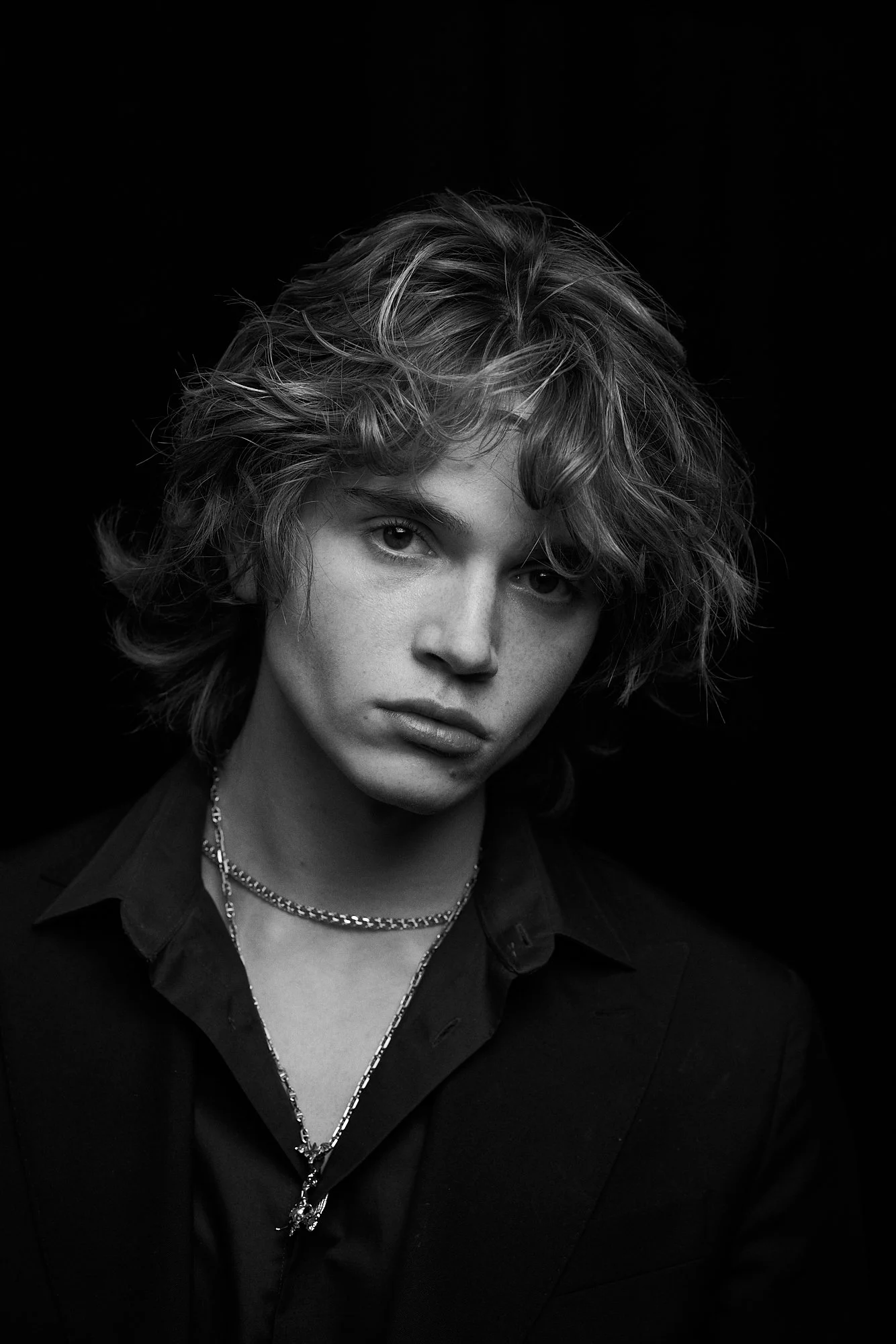 Black and white portrait of a young person with wavy, tousled hair, wearing a dark blazer and layered necklaces, against a dark background.