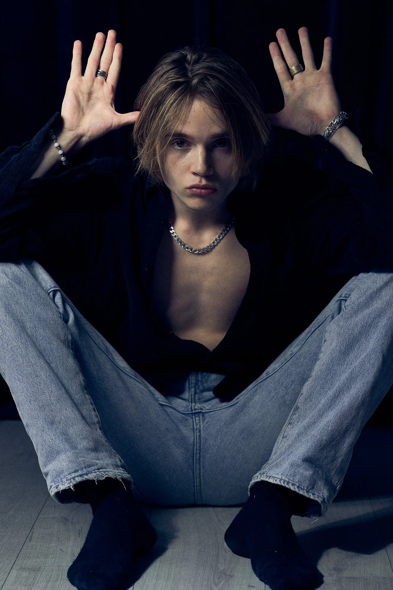 A young woman with short blonde hair sitting cross-legged on the floor, wearing a black shirt, light-wash jeans, black socks, silver jewelry, and looking directly at the camera with hands raised near her head.