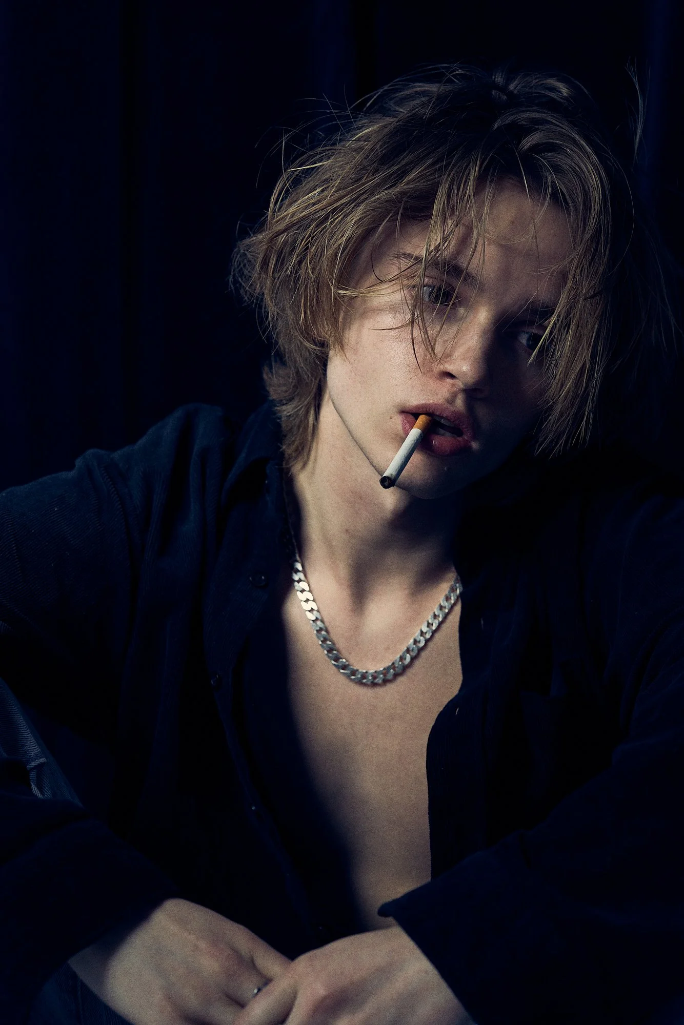 A young man with messy blonde hair, wearing a black shirt, silver chain necklace, and holding a cigarette in his mouth.