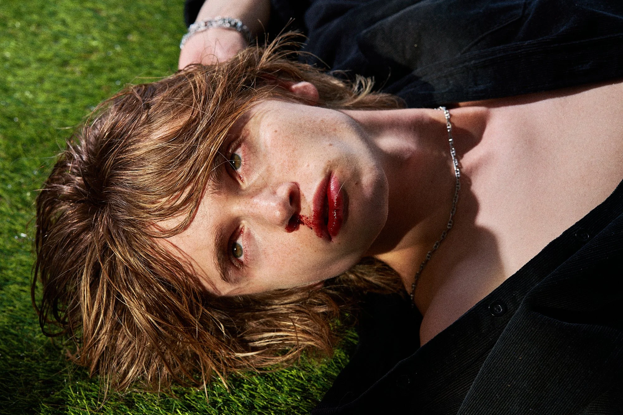 A woman with short wavy hair lying on grass, with blood on her nose and lips, wearing a black shirt and silver necklace.
