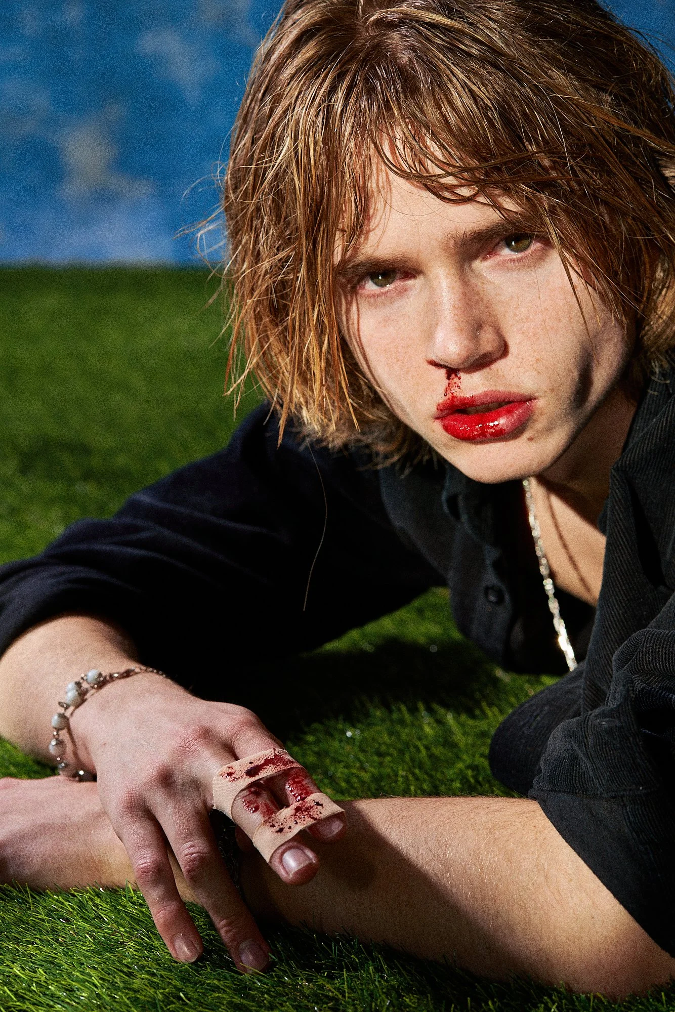 A young woman with short, wet hair, lying on green grass, with blood on her lips and a newborn finger wrapped in blood-stained bandages. She has a serious expression and a nosebleed.