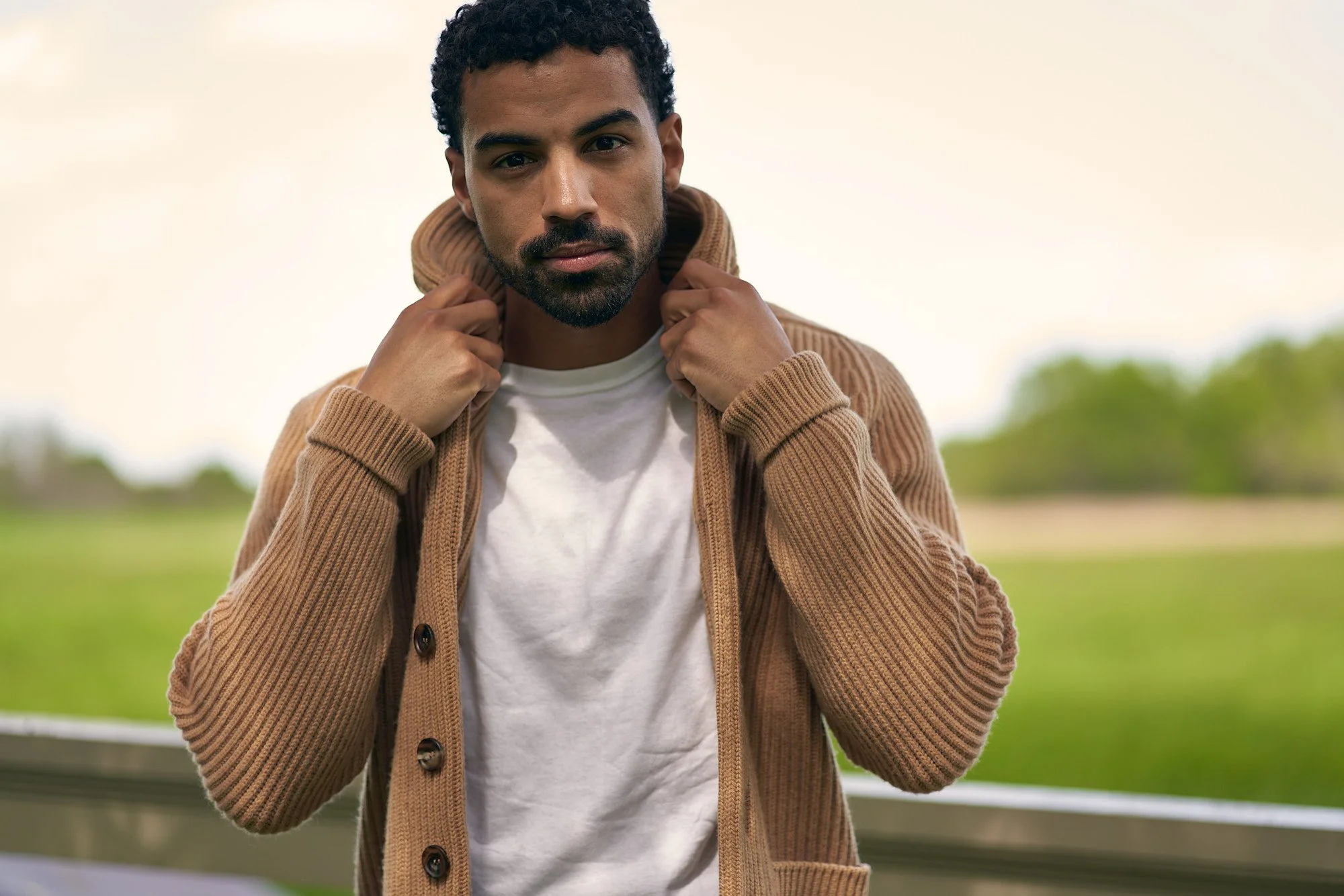 A man with curly hair and a beard wearing a brown cardigan over a white T-shirt, holding the collar of his cardigan, standing outdoors with a blurred green landscape in the background.