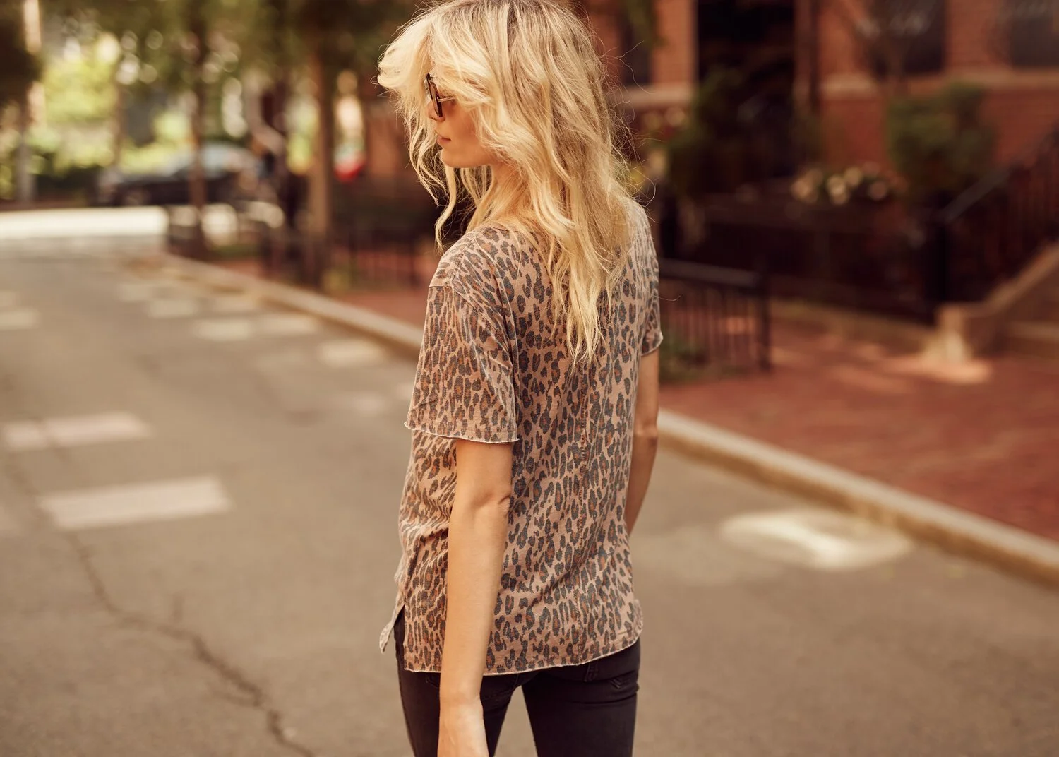A woman with blonde, wavy hair and glasses standing on a city street, wearing a leopard print shirt.