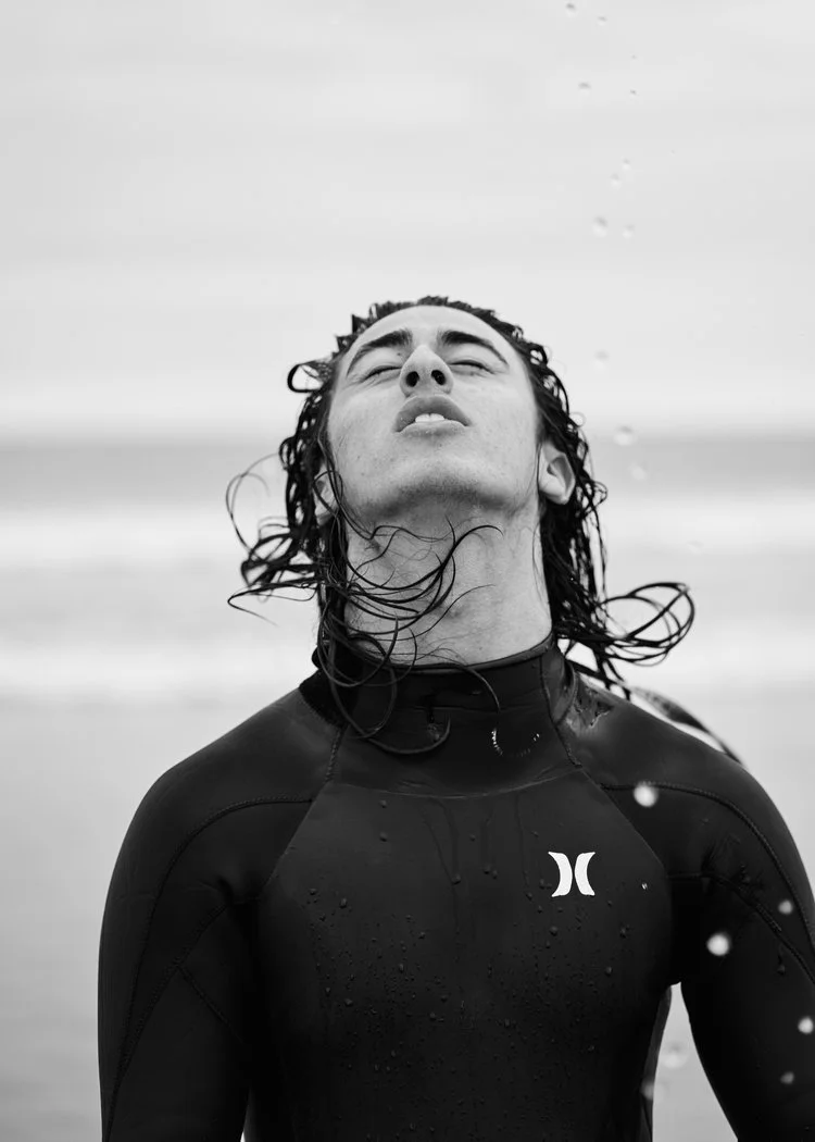 A person with wet hair wearing a wetsuit, standing on a beach with water droplets around, eyes closed, face uplifted.