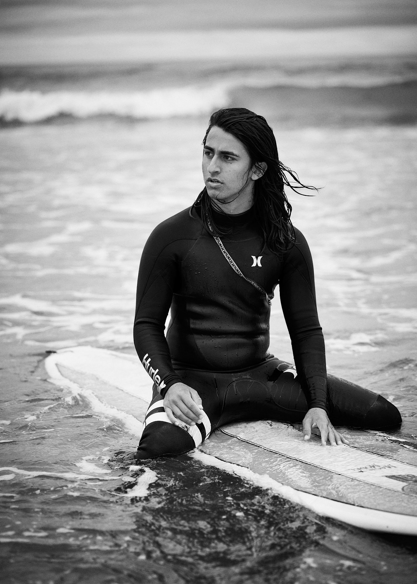 A young man in a wetsuit sitting on a paddleboard in the ocean, looking away, with waves in the background.