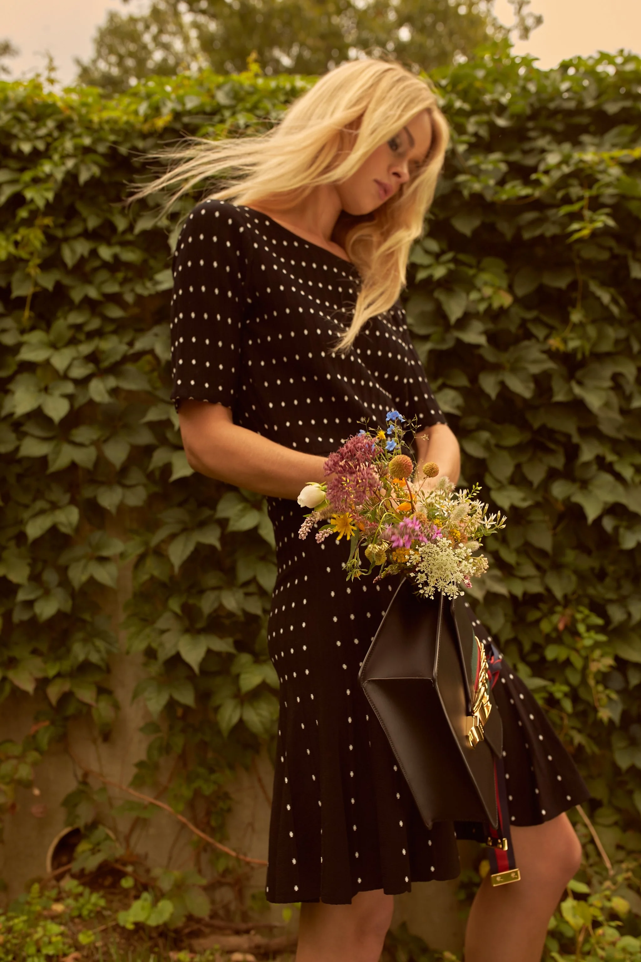 A woman with long blonde hair wearing a black dress with white polka dots holds a bouquet of colorful flowers in a black handbag, standing outdoors in front of a leafy green hedge.