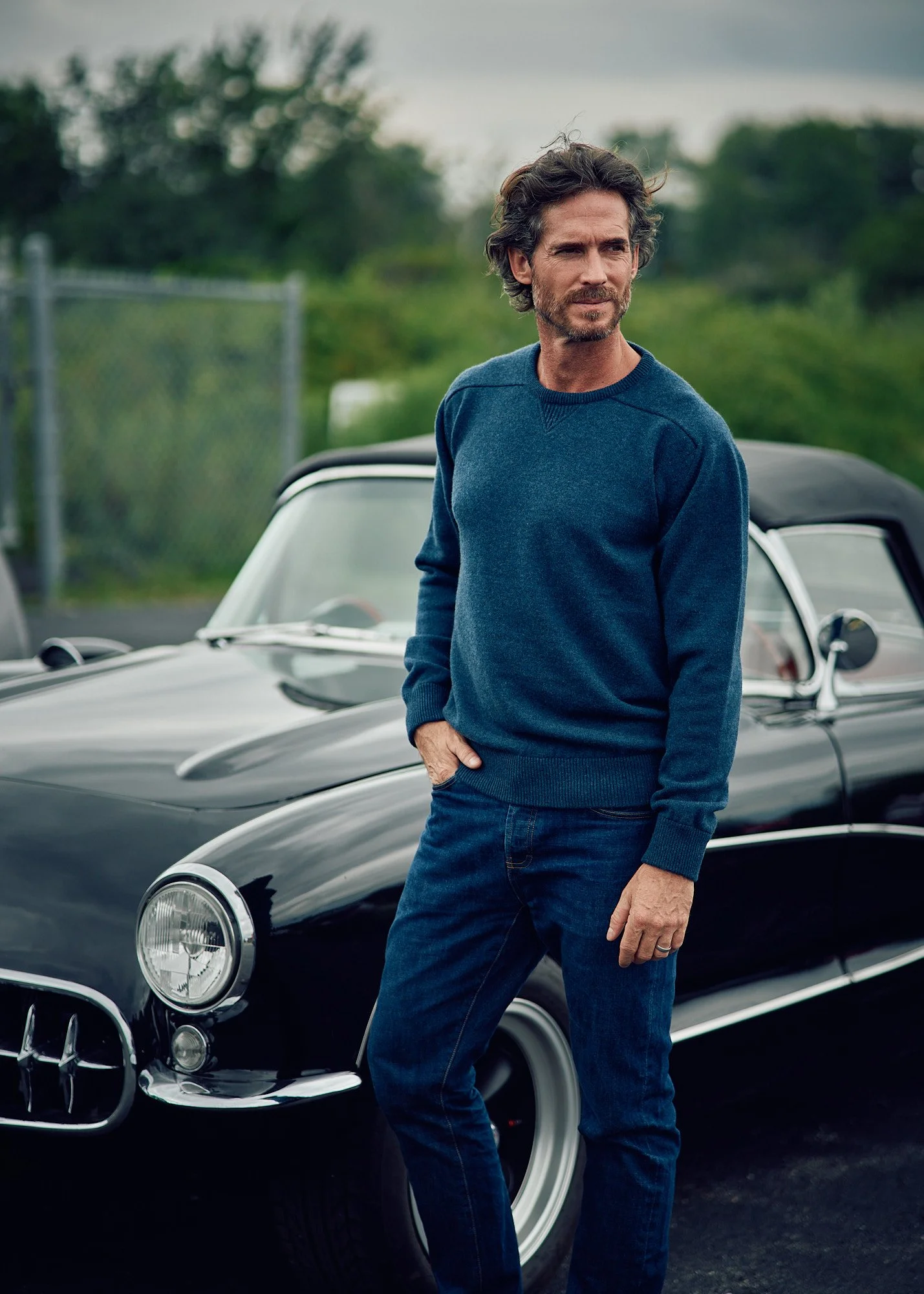 A man with wavy dark hair and a beard standing outdoors near a classic black convertible car. The man is wearing a blue sweater and jeans, with one hand in his pocket. The background features greenery and a cloudy sky.