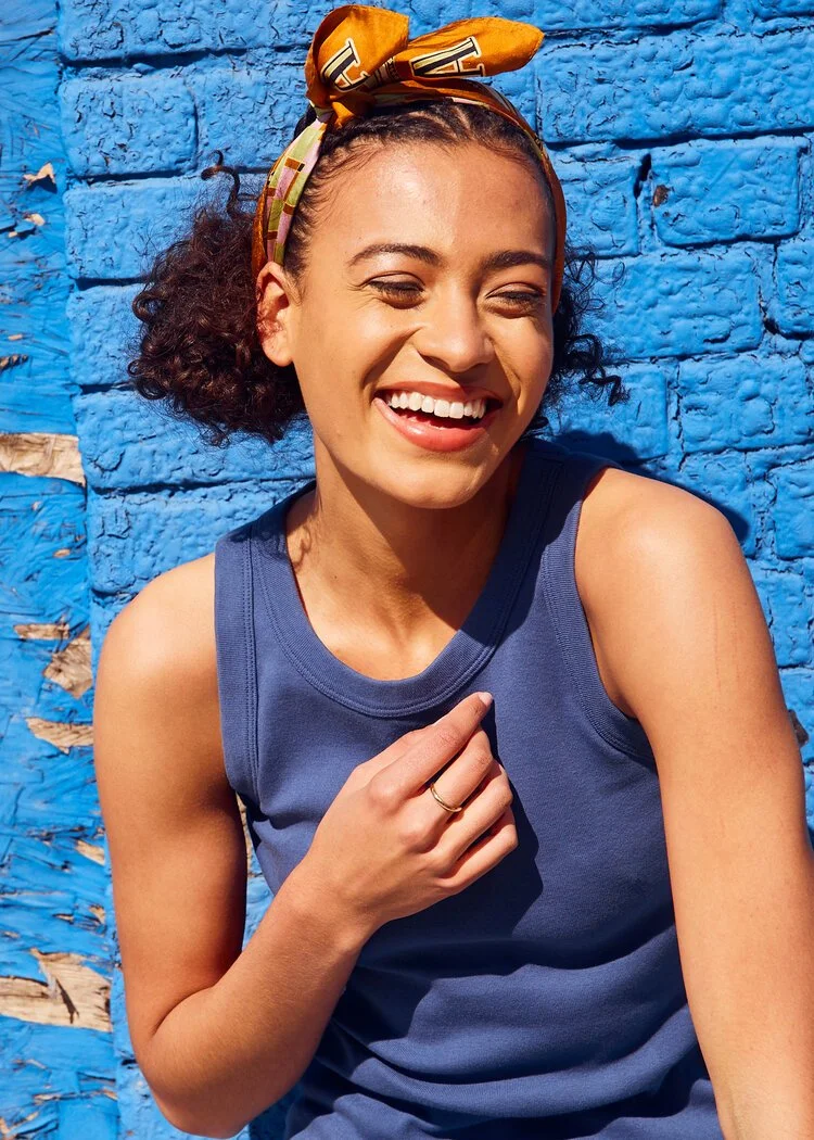 A woman with curly hair, wearing a yellow headscarf, smiling and laughing while sitting against a bright blue wall.