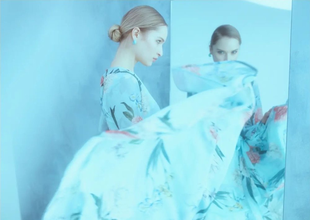 A woman wearing a floral kimono looks at her reflection in a mirror, with her face partially obscured by a flowing piece of fabric she holds.
