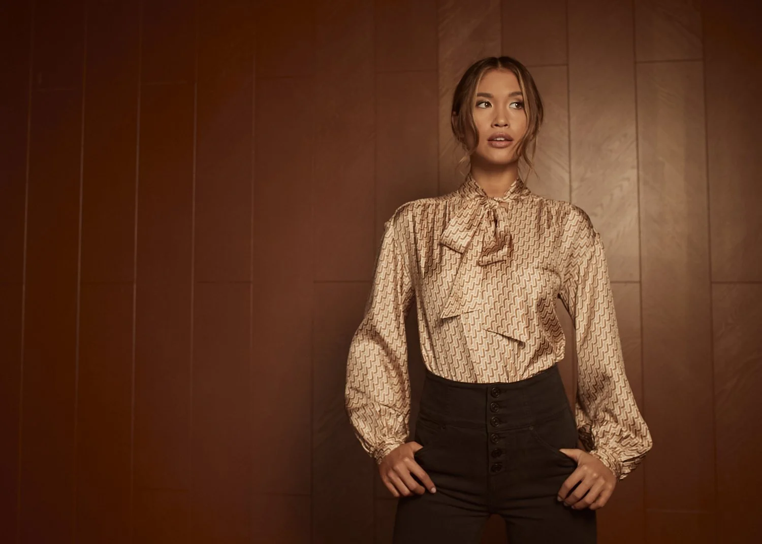 A woman standing against a wooden paneled wall, wearing a patterned blouse with a bow tie at the collar and high-waisted black pants.