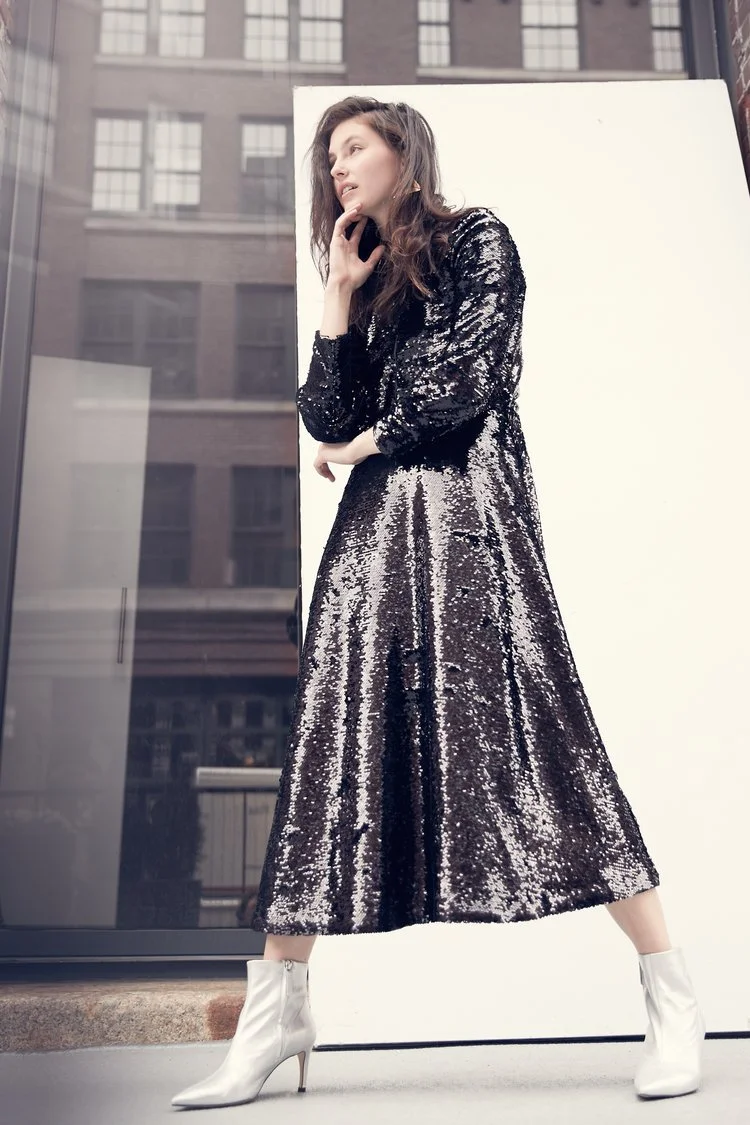 A woman in a black sequined long-sleeve dress and white ankle boots posing outside next to a white background. Urban buildings are reflected in the glass behind her.