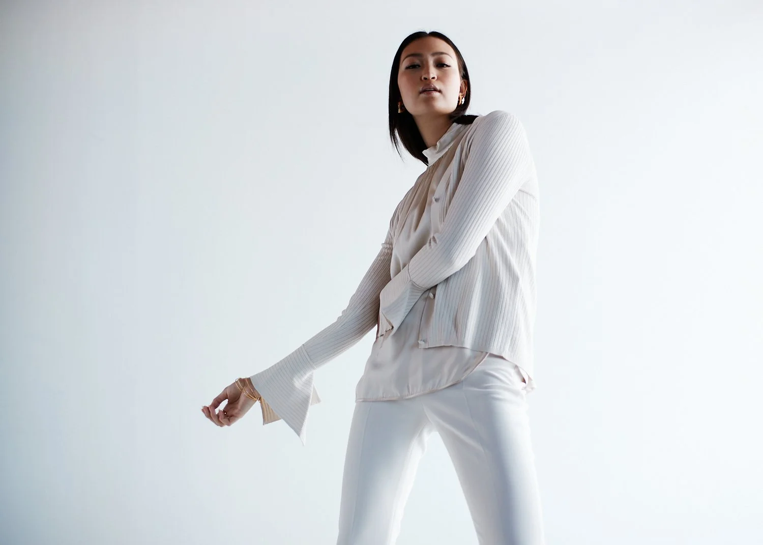 A woman in a cream-colored blouse and white pants standing against a plain background.