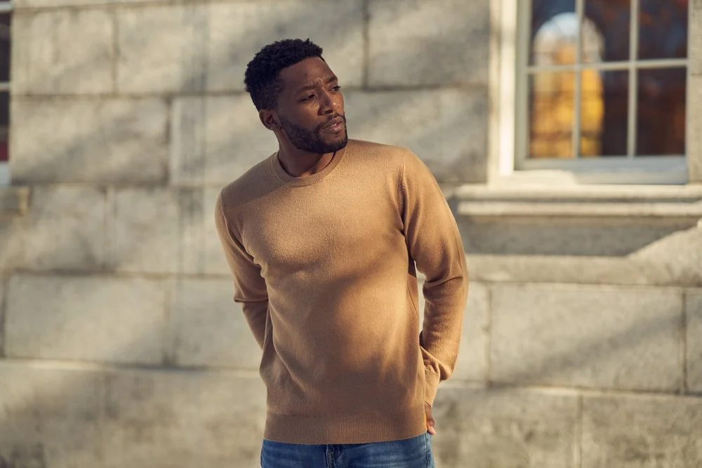 A man with short curly hair and a beard stands outdoors against a stone building with a window, wearing a tan sweater and jeans, looking thoughtful.
