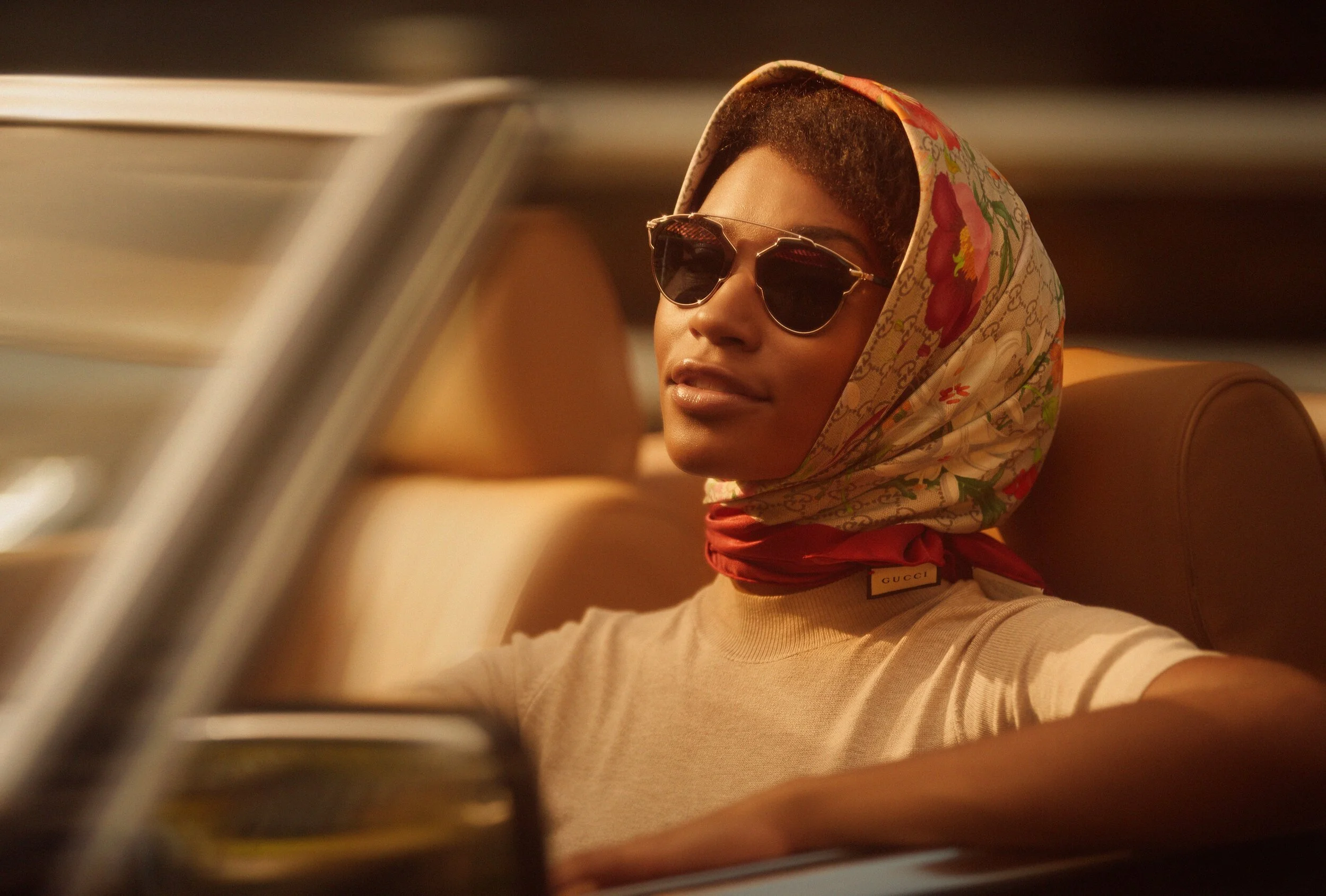 Woman wearing sunglasses and a floral headscarf sitting in a vintage convertible car.