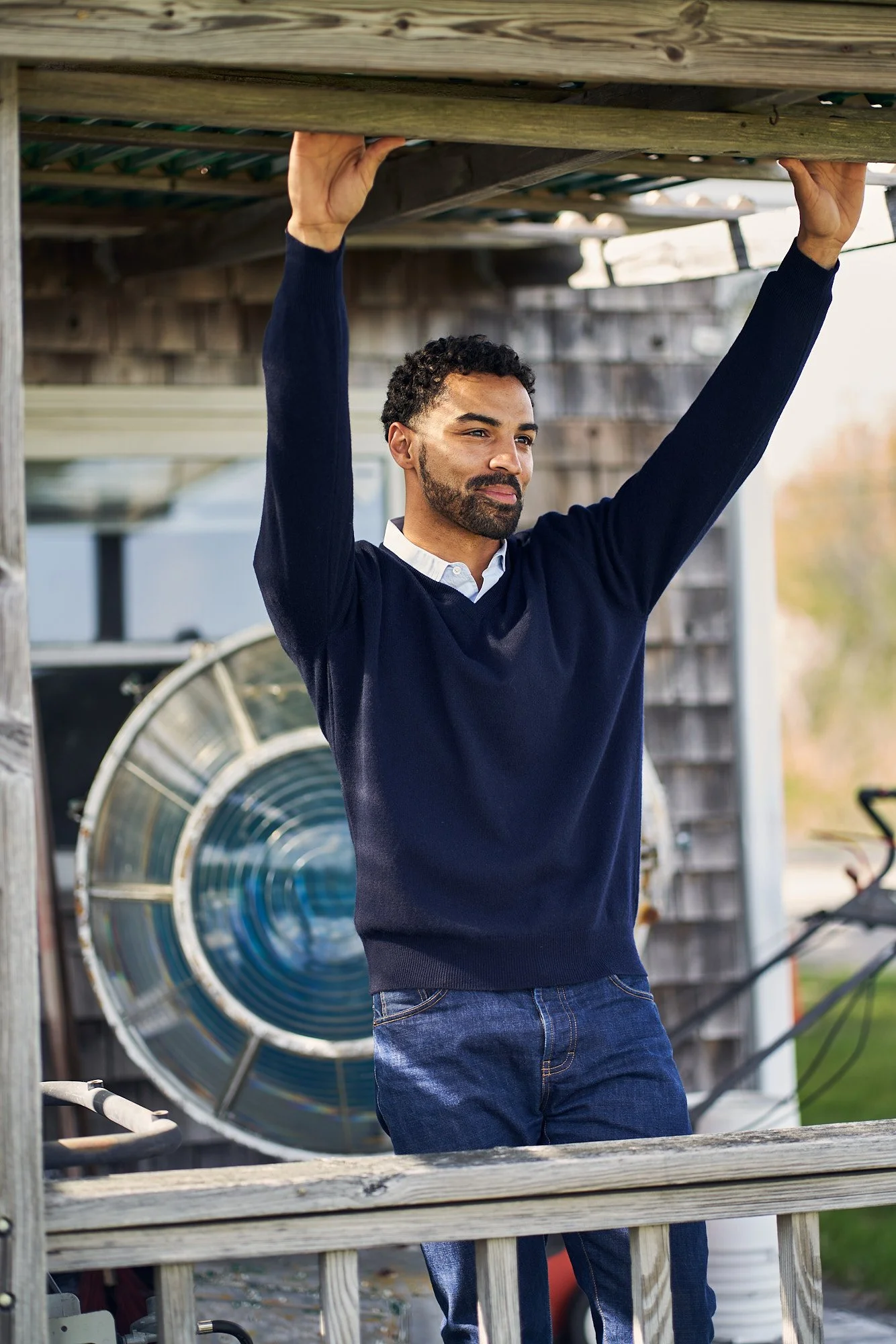 A man with a beard and curly hair wearing a navy blue sweater and jeans, standing on a wooden porch, holding an overhead beam with both hands, smiling slightly, with a garden hose reel in the background.