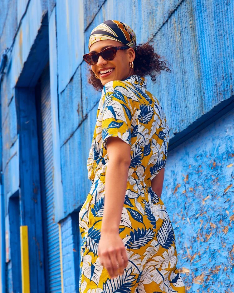 A woman wearing sunglasses, a patterned headscarf, and a colorful floral jumpsuit stands against a vibrant blue textured wall, smiling and looking back.