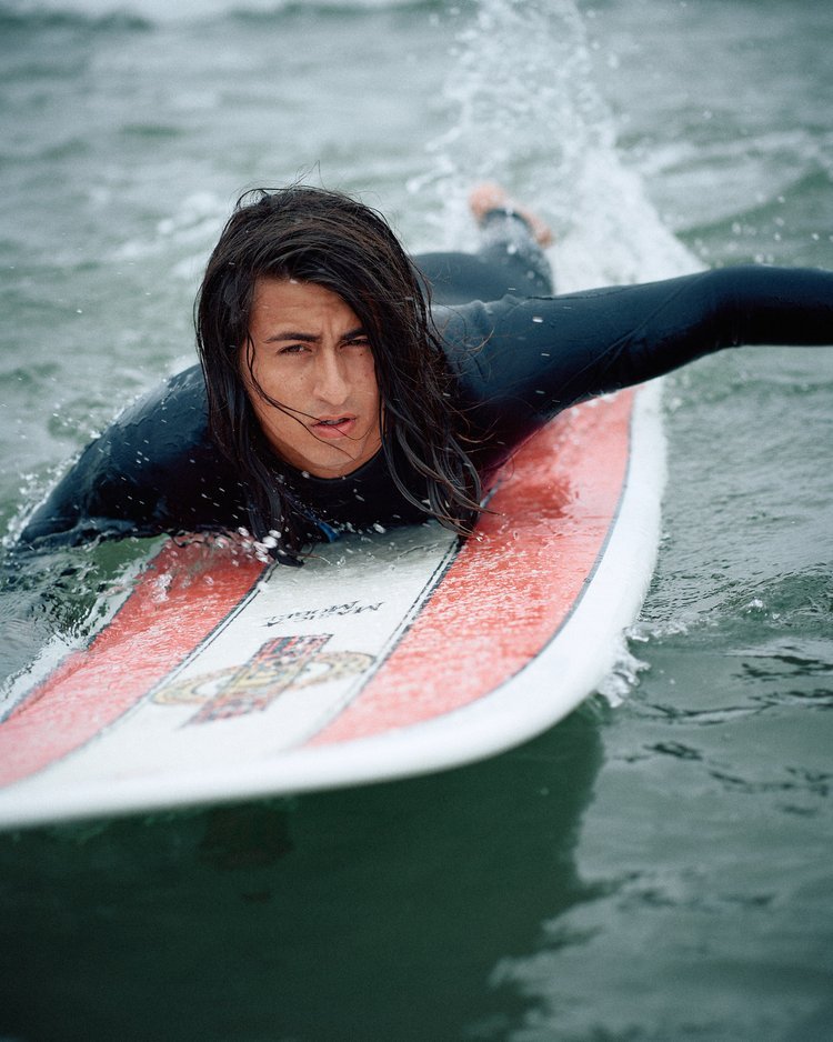 Young woman with wet hair, lying on a surfboard in the water, wearing a black wetsuit, on a cloudy day.
