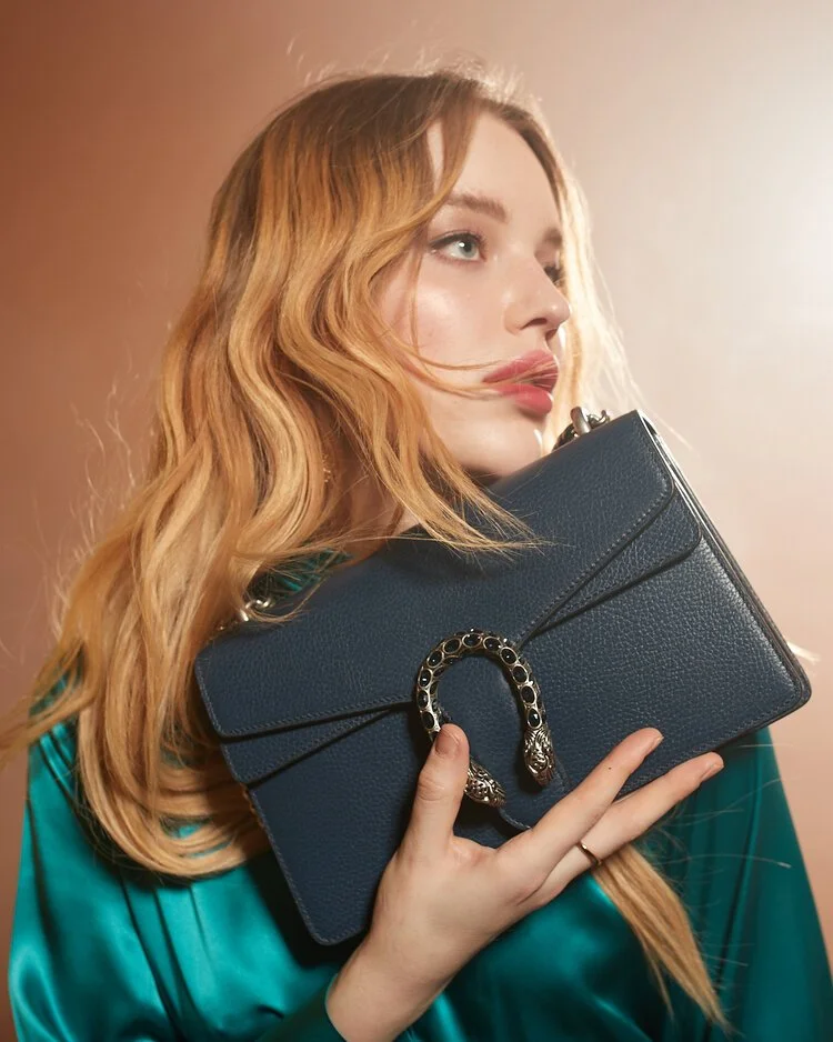 A woman with long, wavy blonde hair holding a navy blue designer clutch purse with a decorative snake-shaped clasp.