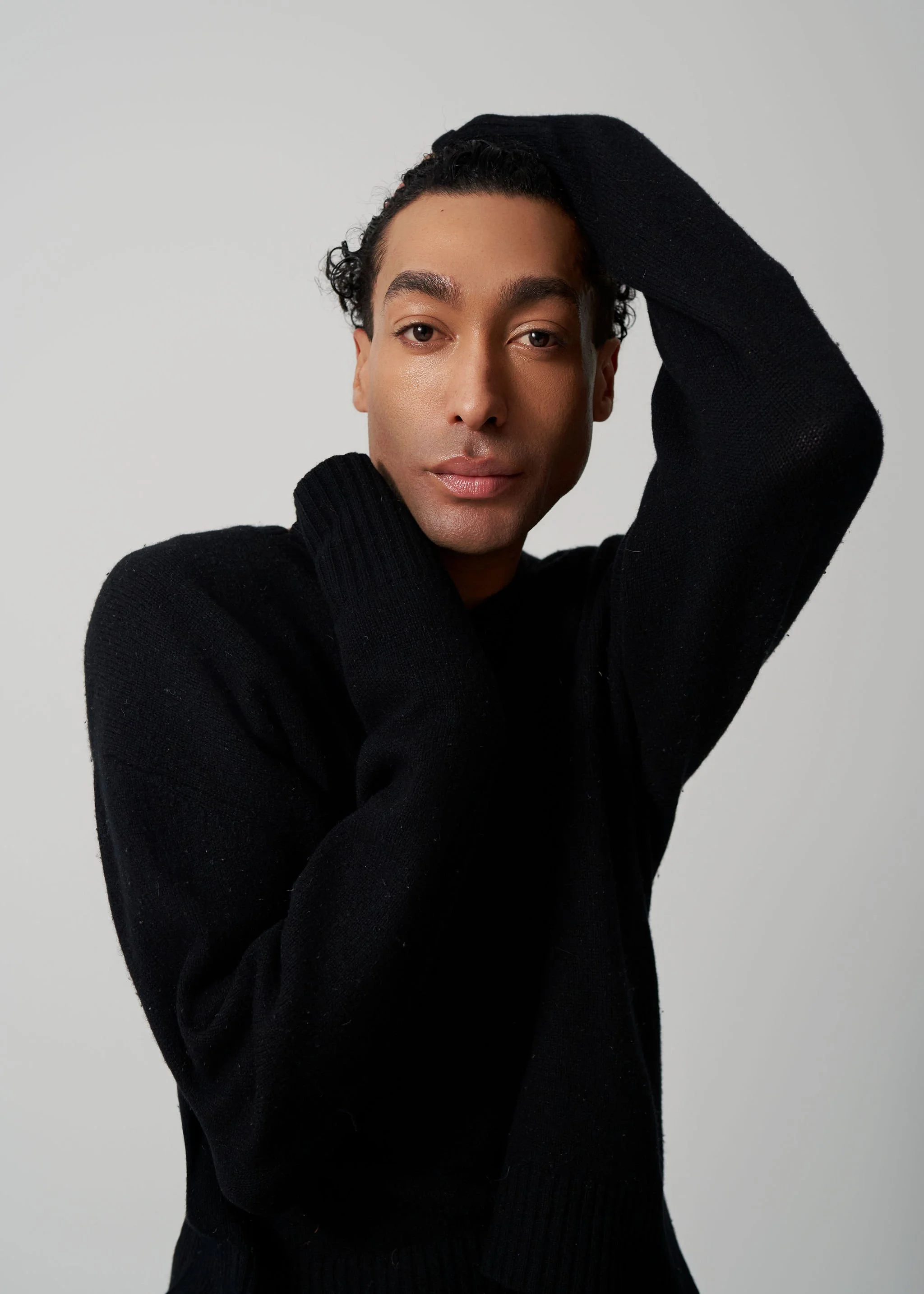 A person with medium skin tone and short, curly dark hair, wearing a black sweater, poses with one arm raised behind their head and the other hand touching their neck, against a light gray background.