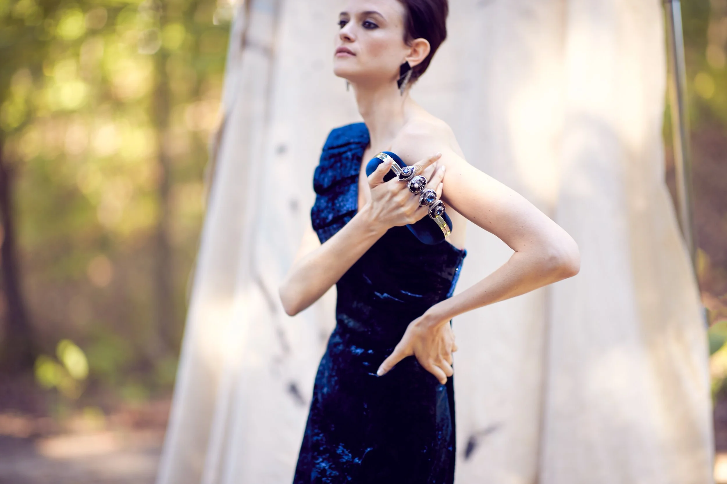 A woman in a dark blue velvet dress with ruffled shoulder detail standing outdoors against a white fabric backdrop with trees in the background.