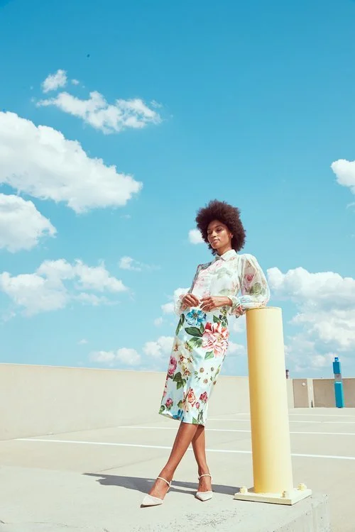 A woman standing on a parking lot under a bright blue sky with scattered clouds, leaning against a yellow parking barrier, wearing a floral dress and high heels.