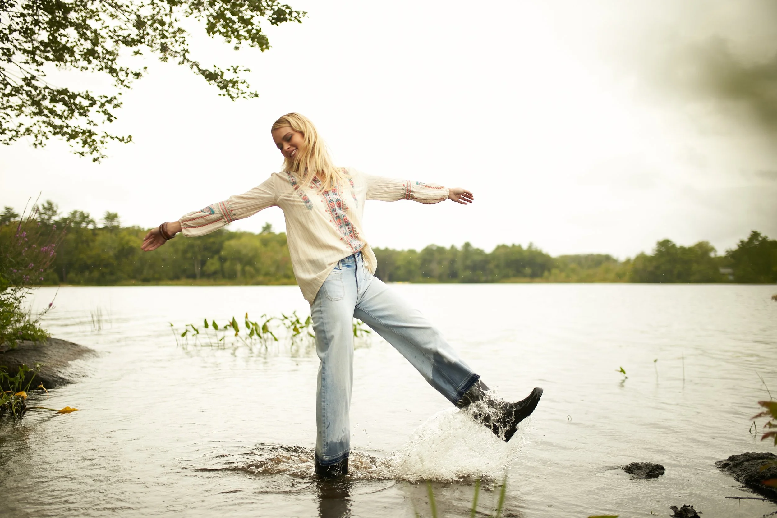 Young woman in a long-sleeve blouse and jeans enjoying herself while kicking water in a lake or river during daytime.