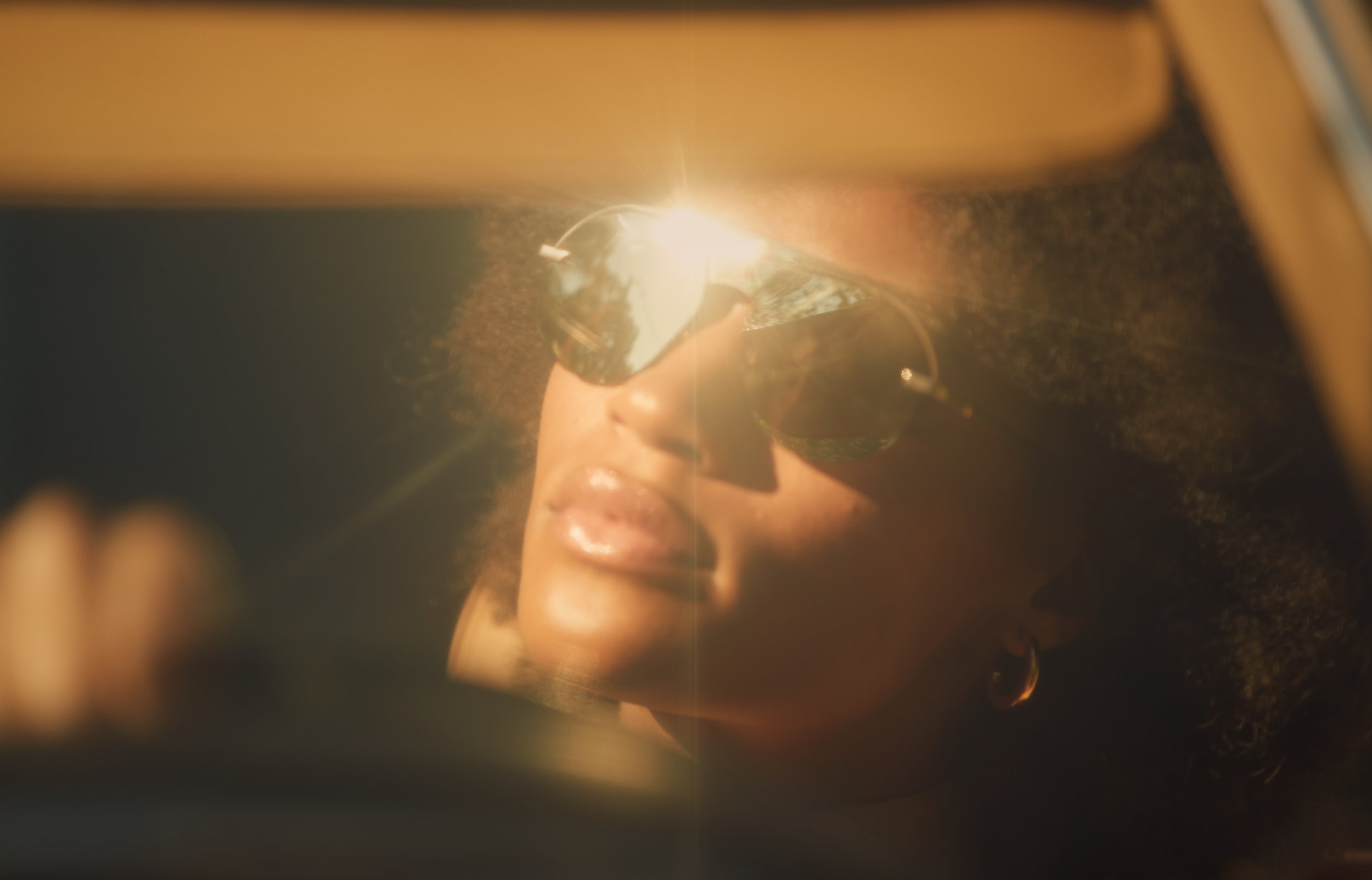 A woman with curly hair is wearing mirrored sunglasses and looking at her reflection, with sunlight reflecting off the sunglasses.