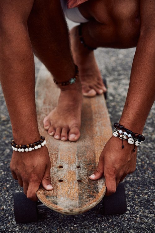 Person bending over, adjusting a skateboard on asphalt, wearing multiple beaded bracelets on both wrists.