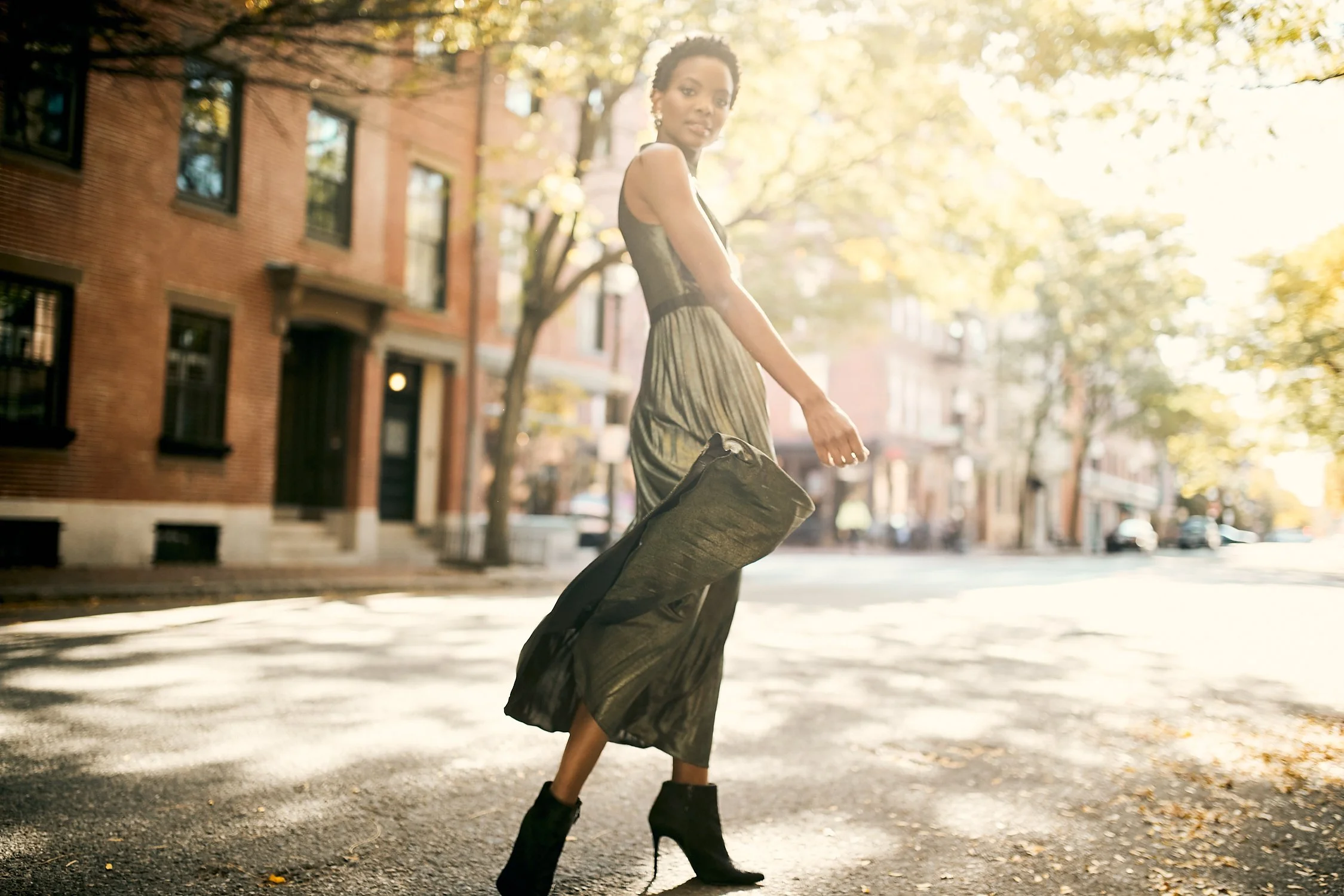 A woman in an elegant dress and high heels walks on a sunlit city street with brick buildings and trees in the background.
