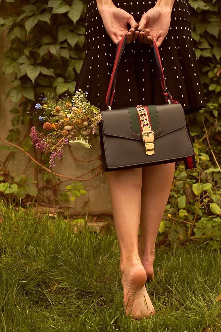 A woman holding a black handbag with a gold lock and striped strap, with a bouquet of colorful wildflowers, standing barefoot on grass in front of green leafy plants.