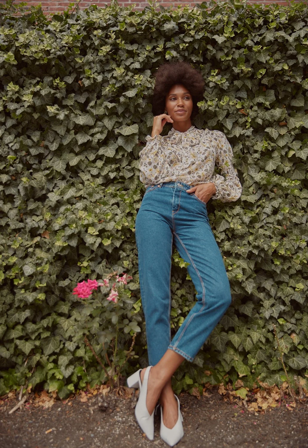 A woman with natural curly hair wearing a floral blouse and blue jeans, standing against a background of dense green ivy and pink flowers.