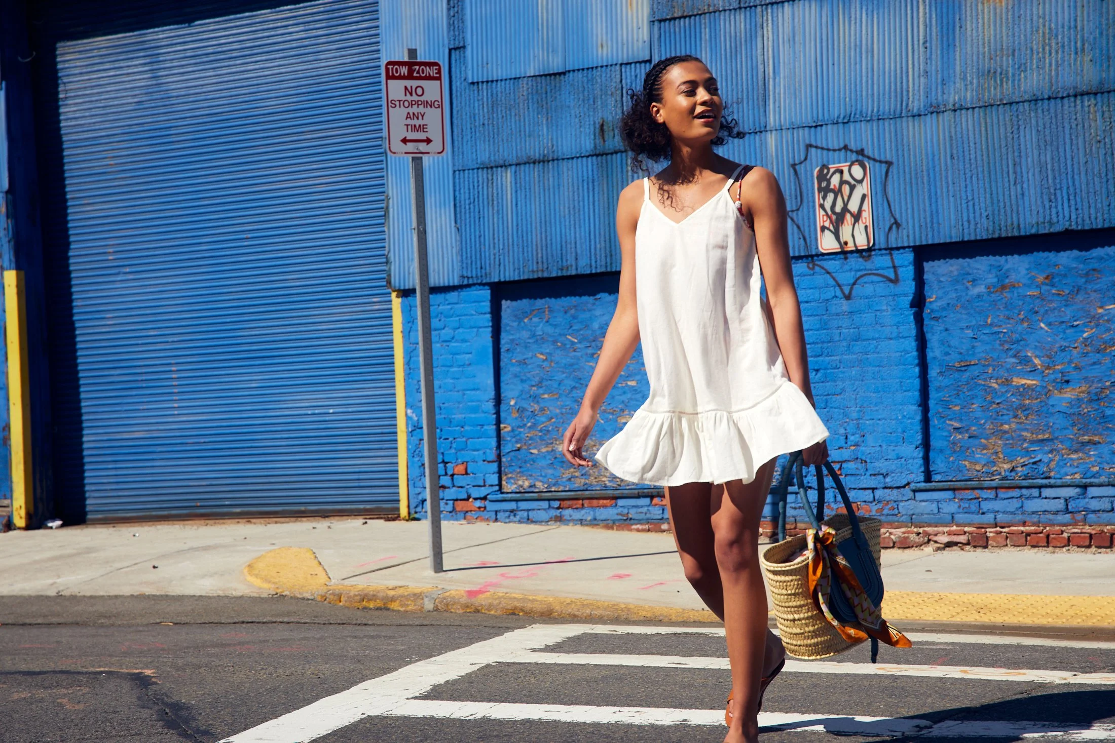 Young woman in a white dress walking across a crosswalk carrying a straw bag with a scarf tied to it, with a blue wall and a 'No Stopping Any Time' sign in the background.