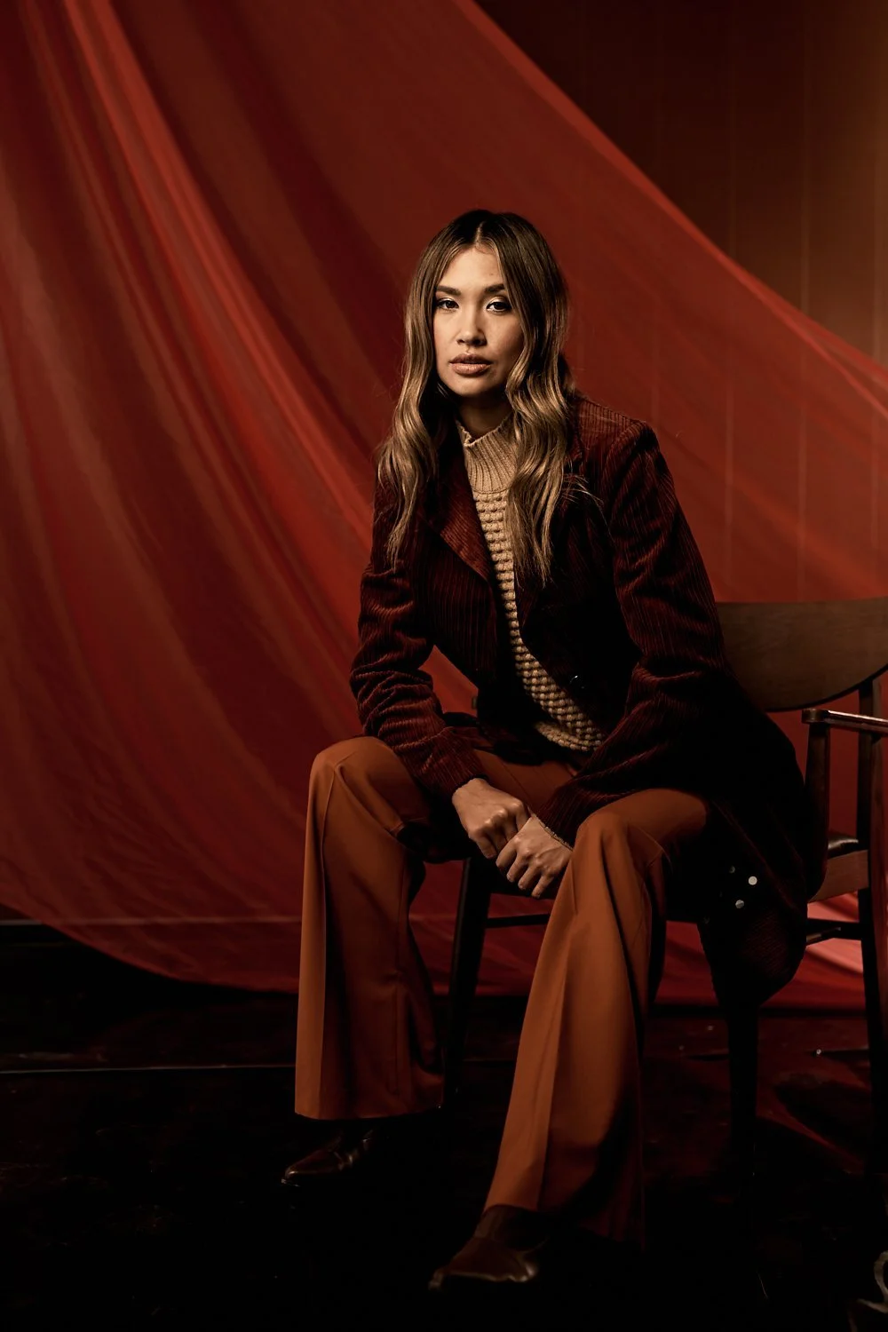 A woman with long wavy hair sitting on a wooden chair against a red fabric backdrop, wearing a burgundy velvet jacket, beige ribbed sweater, and tan wide-leg pants.