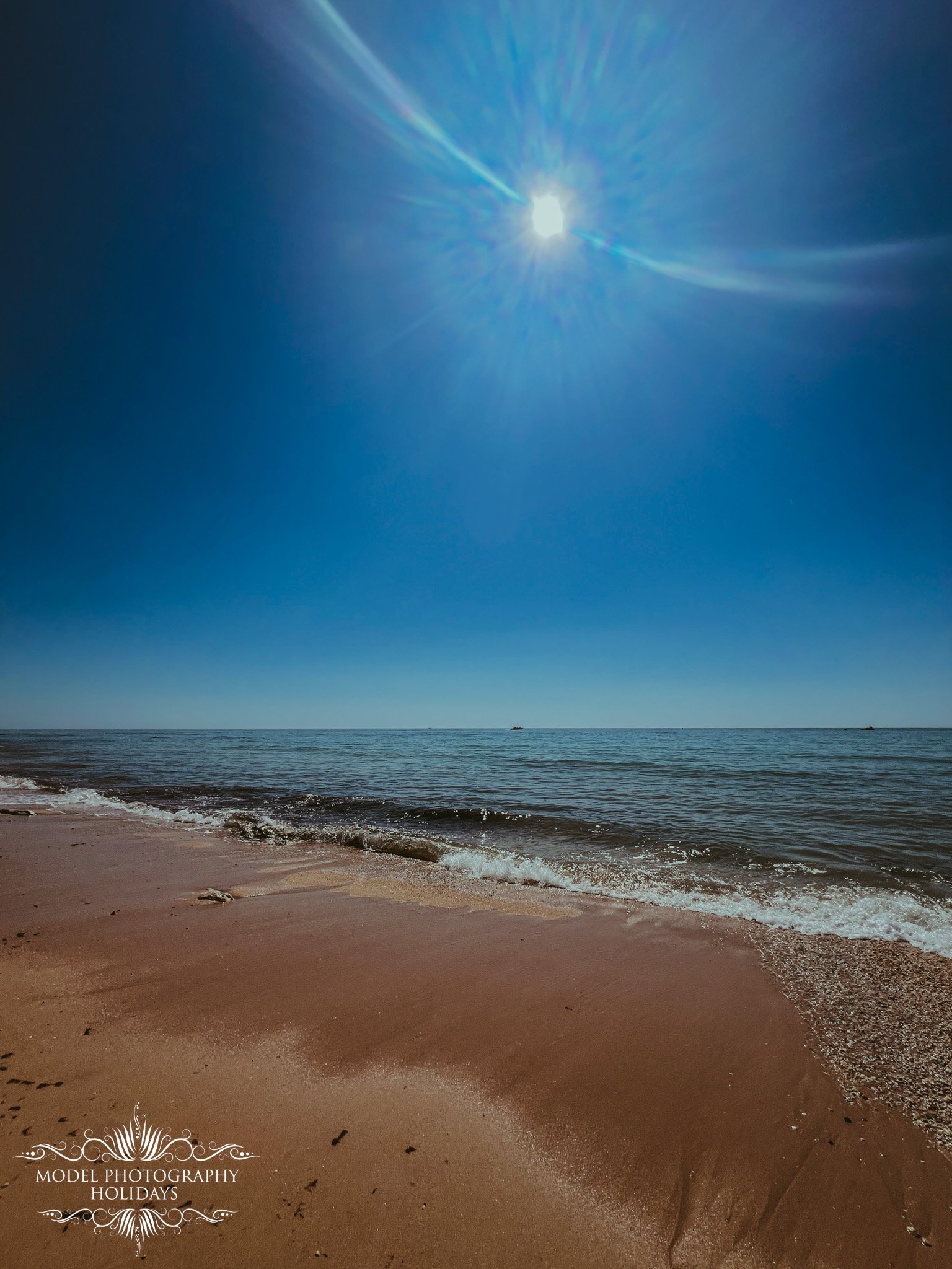 View of a sandy beach with gentle waves, the ocean stretching out to the horizon, and a bright sun shining in a clear blue sky.