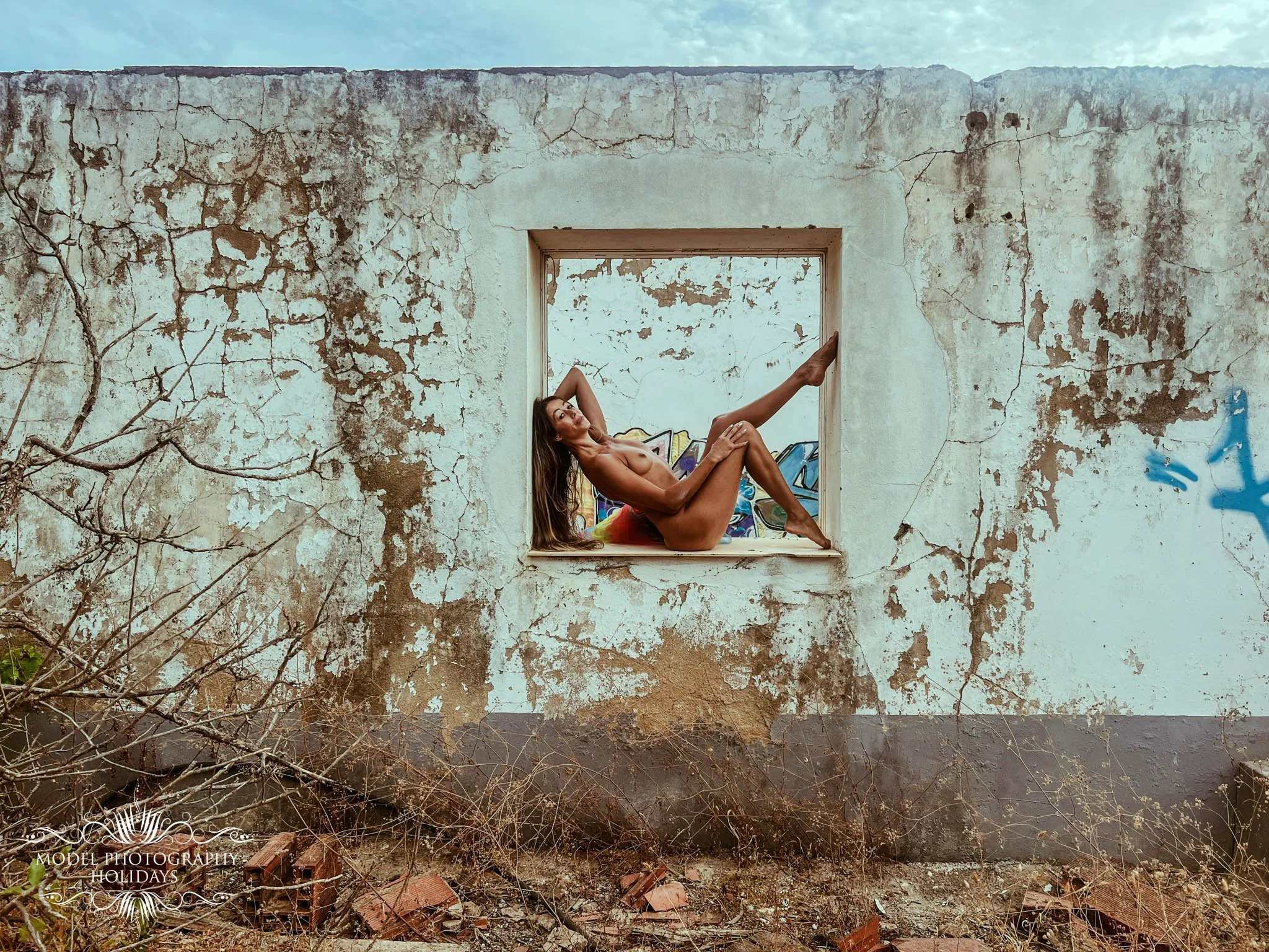 Person with long hair and colorful shorts sitting in an arched window opening of an old, weathered building with peeling paint and cracks, surrounded by dry vegetation.