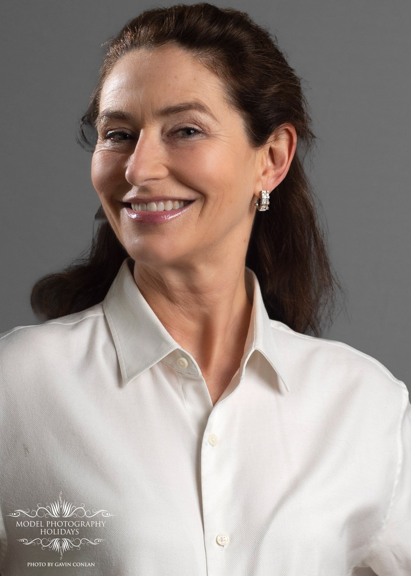 Smiling woman in a white button-up shirt with earrings, against a gray background.