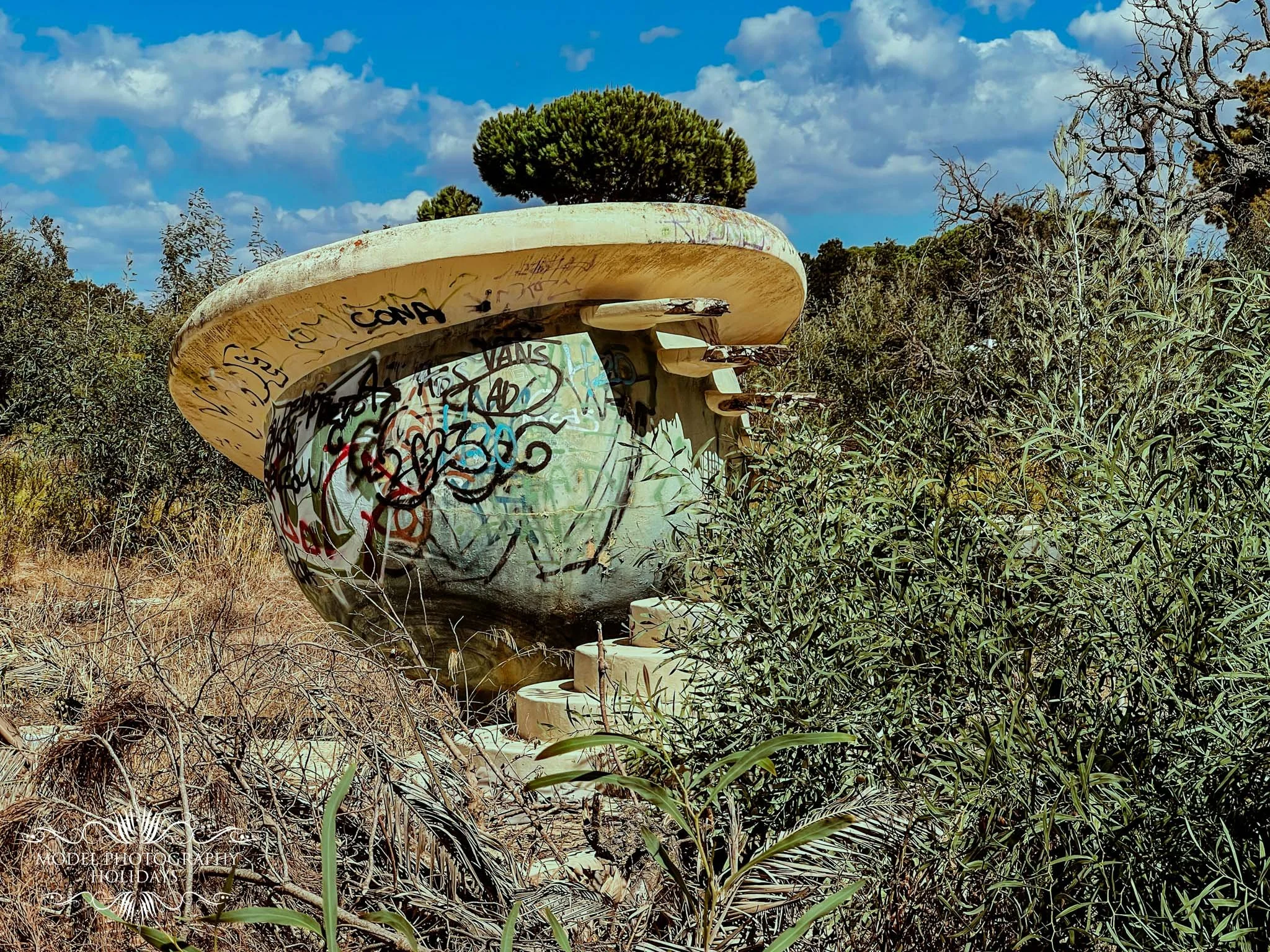 A large boat hull with graffiti on it, partially cut off, surrounded by dry grass and bushes, with a blue sky and scattered clouds in the background.