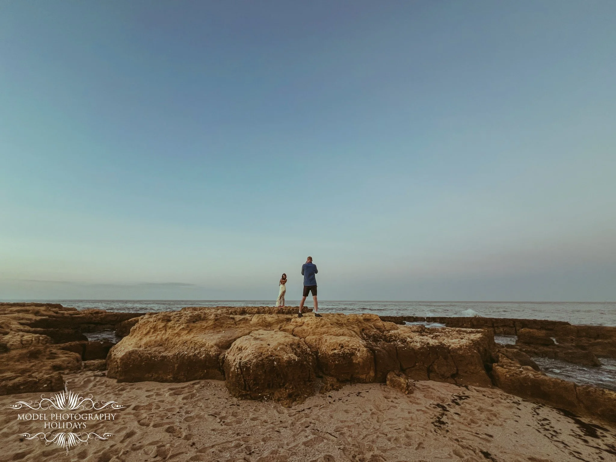 A person and a woman standing on rocks by the ocean, with a man taking a photo of the woman, under a clear blue sky.