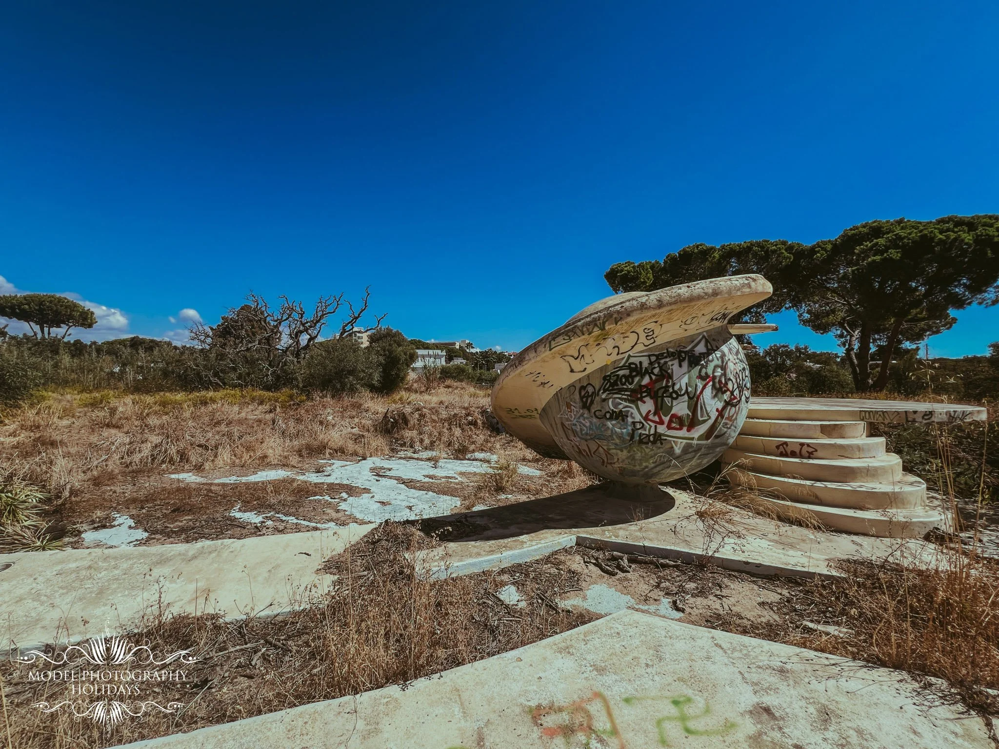 An outdoor scene featuring a large, modern, abstract sculpture covered in graffiti, surrounded by dry grass, trees, and a bright blue sky.